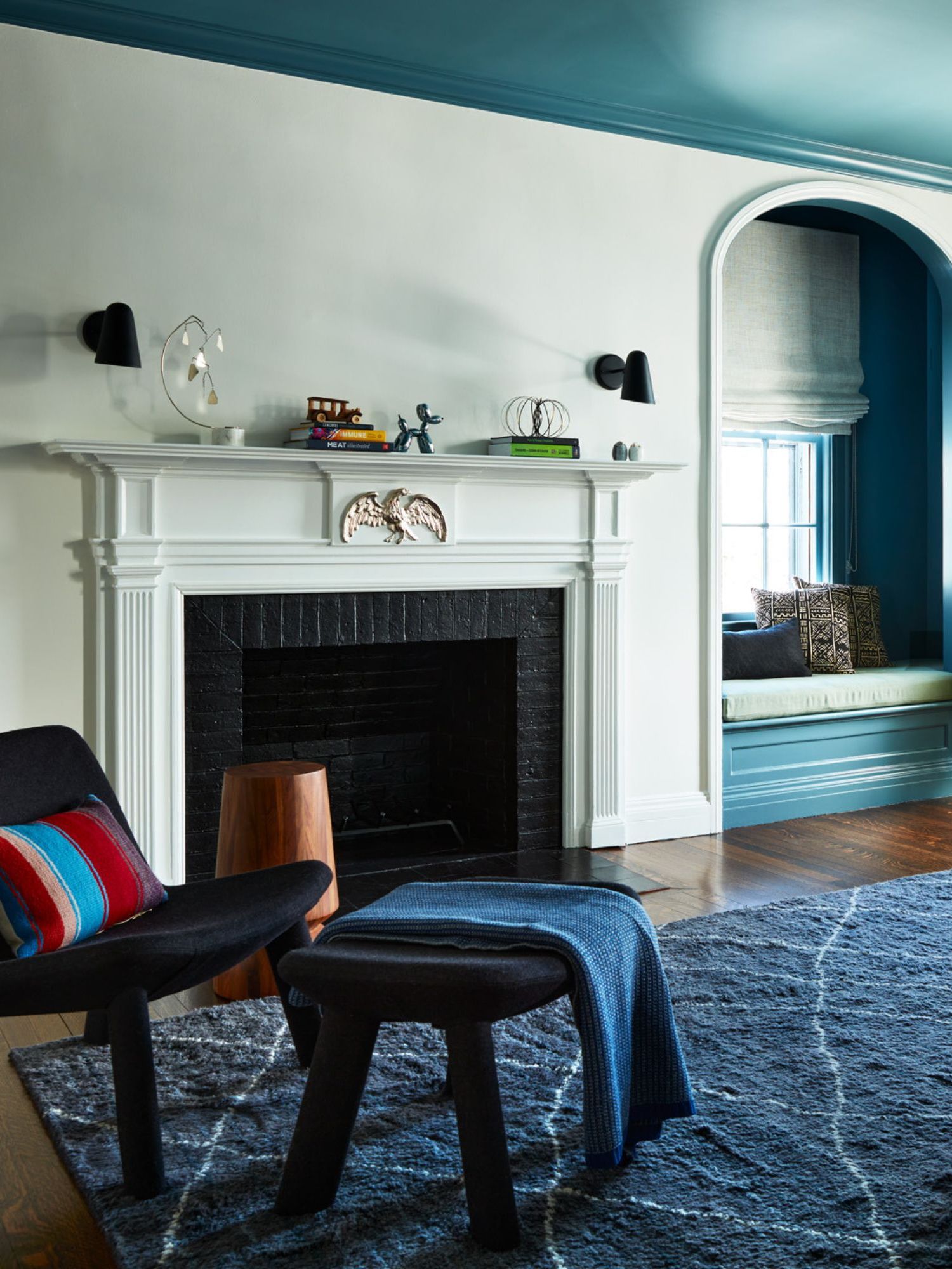 A large bedroom with white walls and a teal ceiling, with an alcove window seat painted teal. A large white fireplace, a navy rug, a dark blue accent chair, and pale blue drapes.