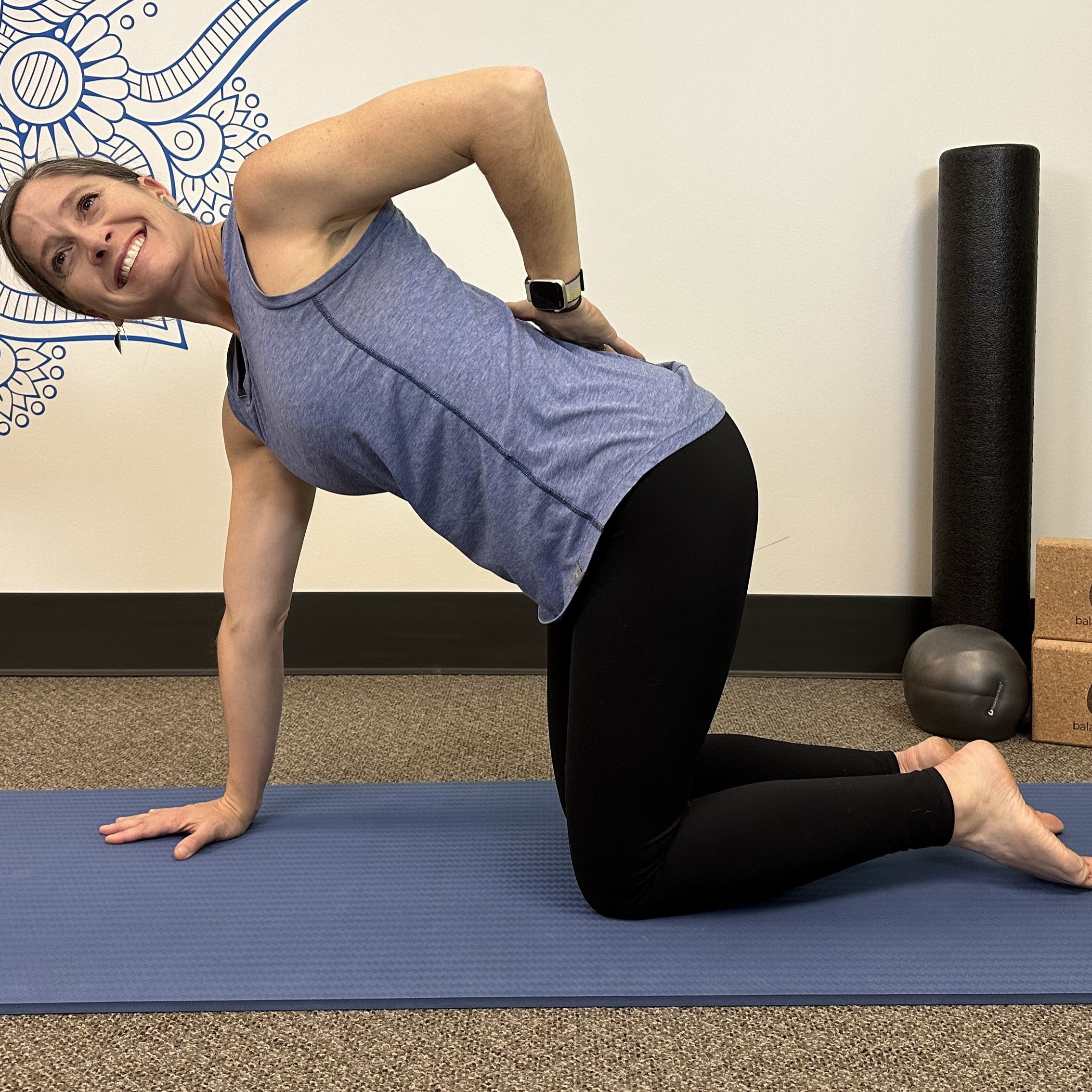 woman in a blue vest and black leggings on a blue exercise mat with physio props behind her. she&#039;s on her hands and knees with one arm on her lower back, chest rotated toward the camera and smiling