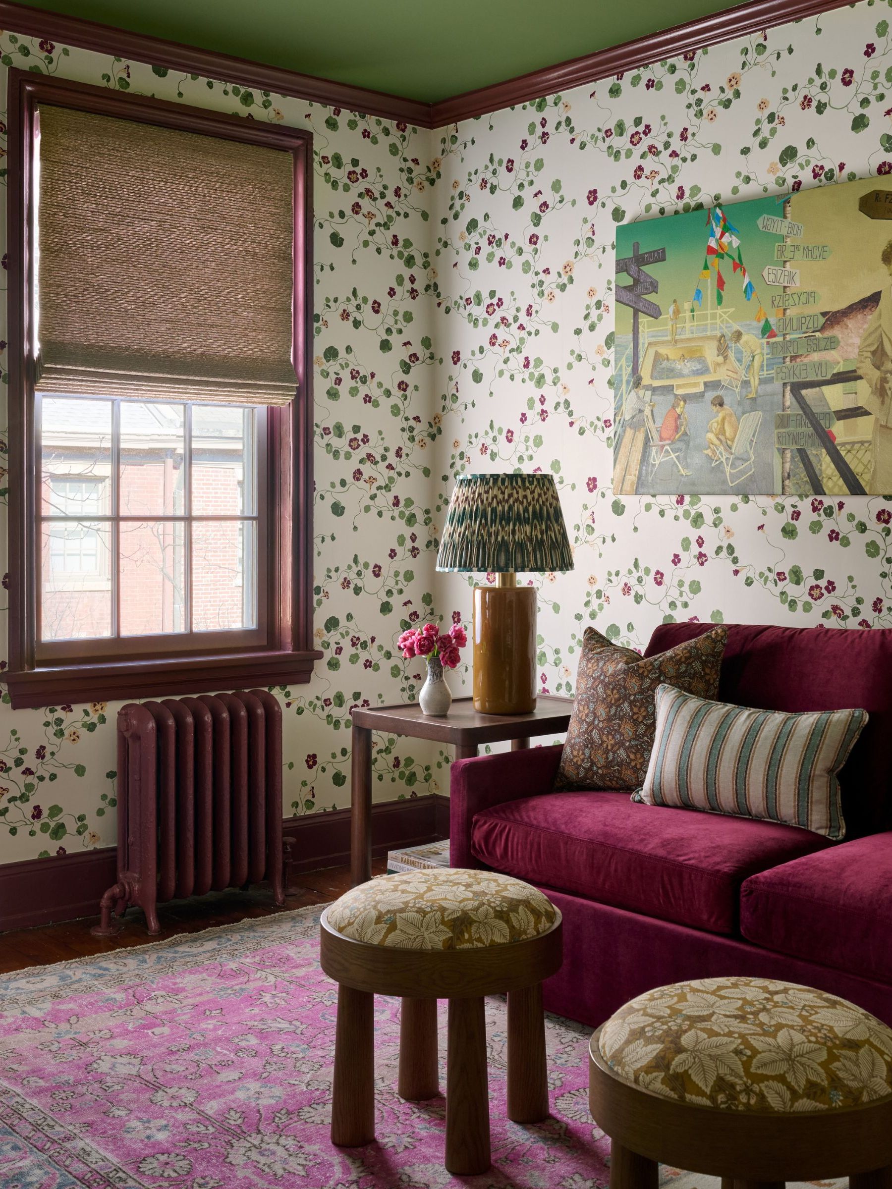 Sitting room corner with floral wallpaper, a burgundy velvet sofa, a pink patterned rug, and two small round wooden stools.