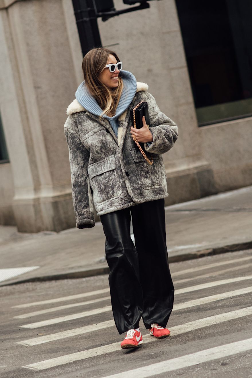 a fashion week attendee crossing the street in blue hoodie, grey jacket, leather pants, and red sneakers