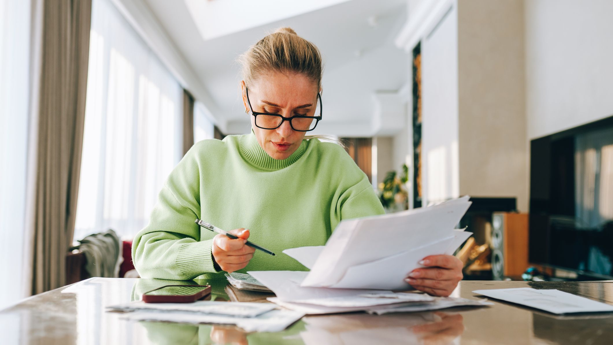 A woman in glasses concentrating on paperwork, holding documents and a pen while budgeting.