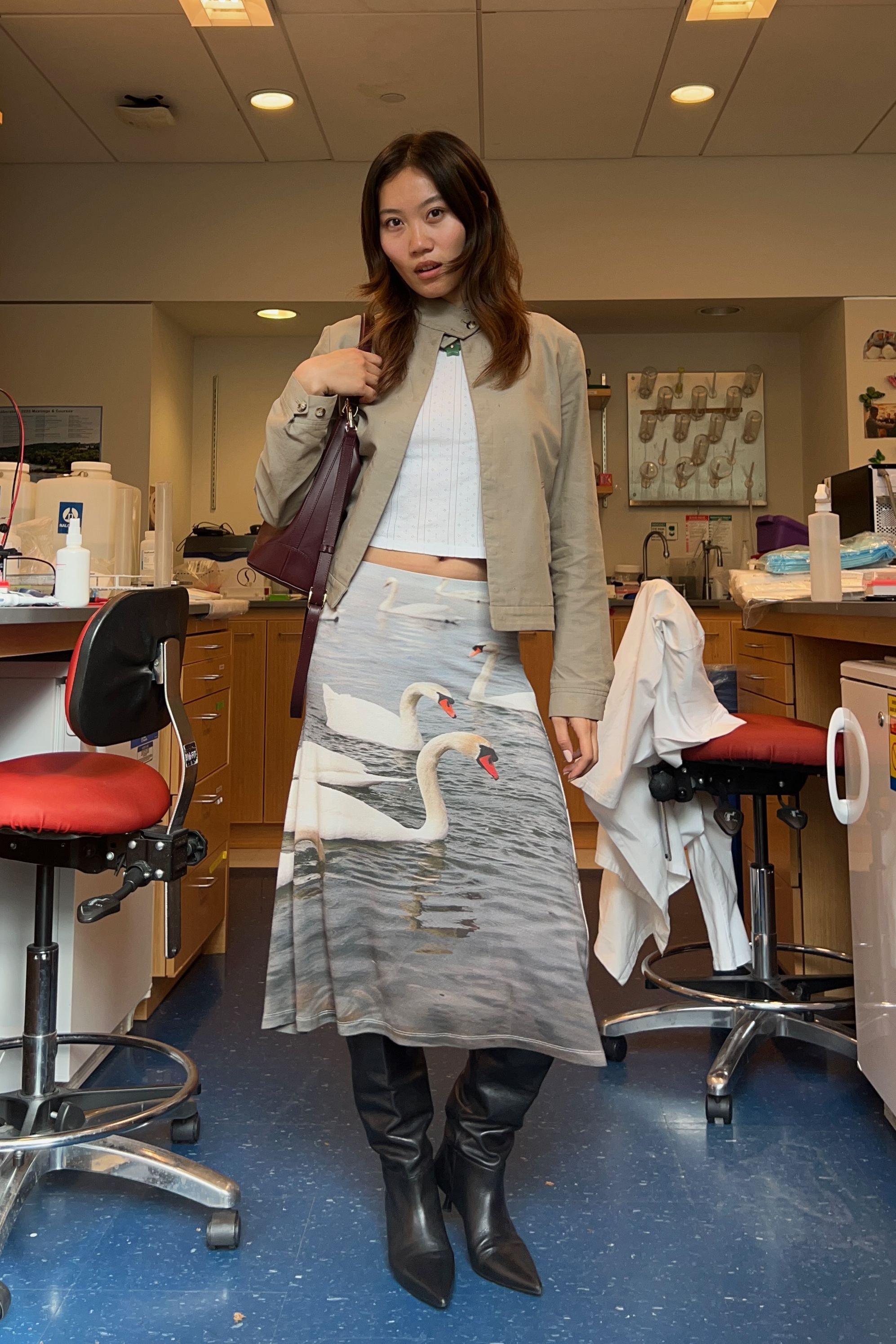 Vivian Li wearing a swan print skirt, white tee, tan jacket, black boots, and red leather bag in a science lab.