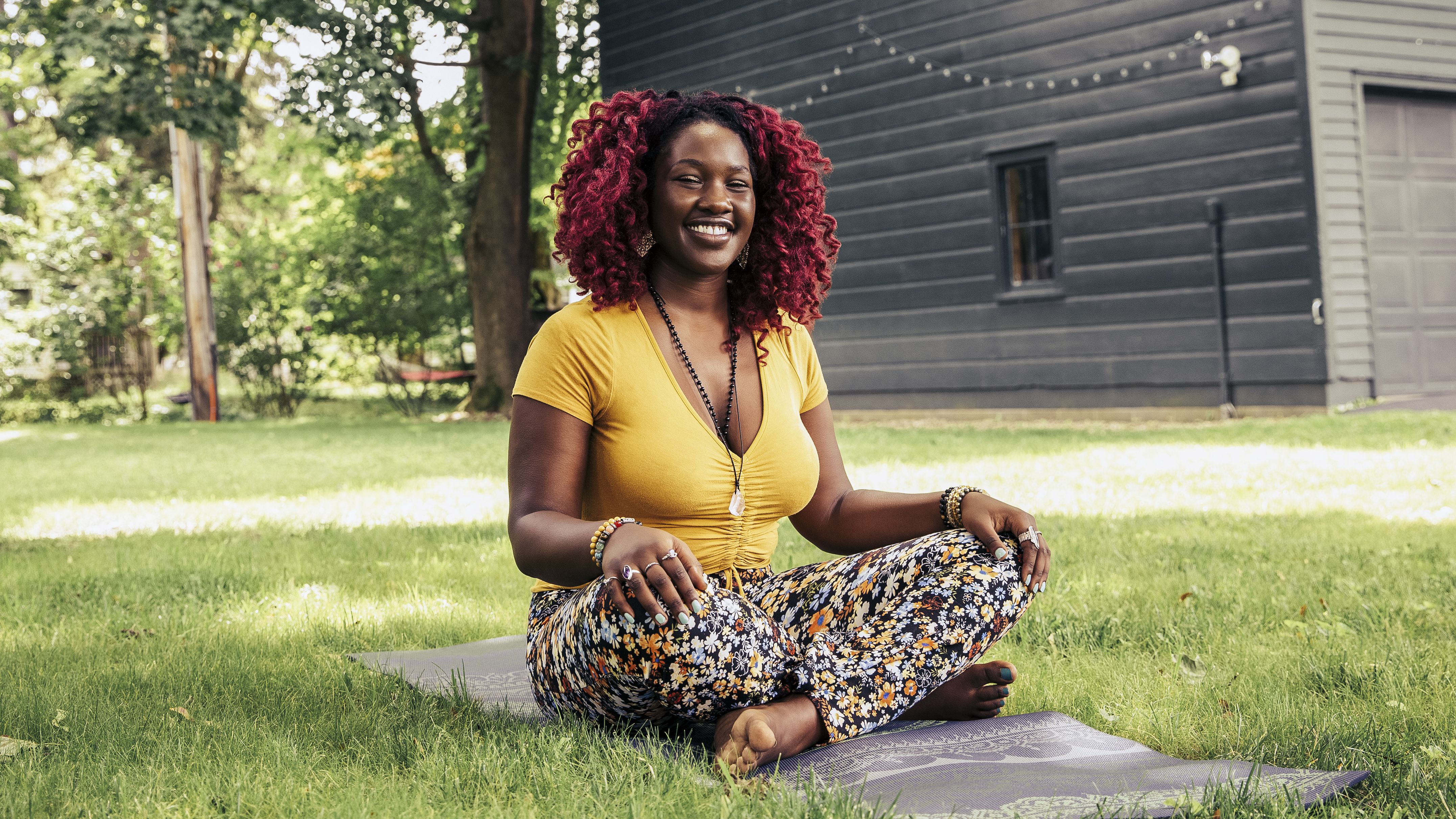 A woman sits cross-legged outside on a yoga mat. Behind her we see a large expanse of healthy-looking grass and the side of a house with a garage, which is decorated with fairy lights.