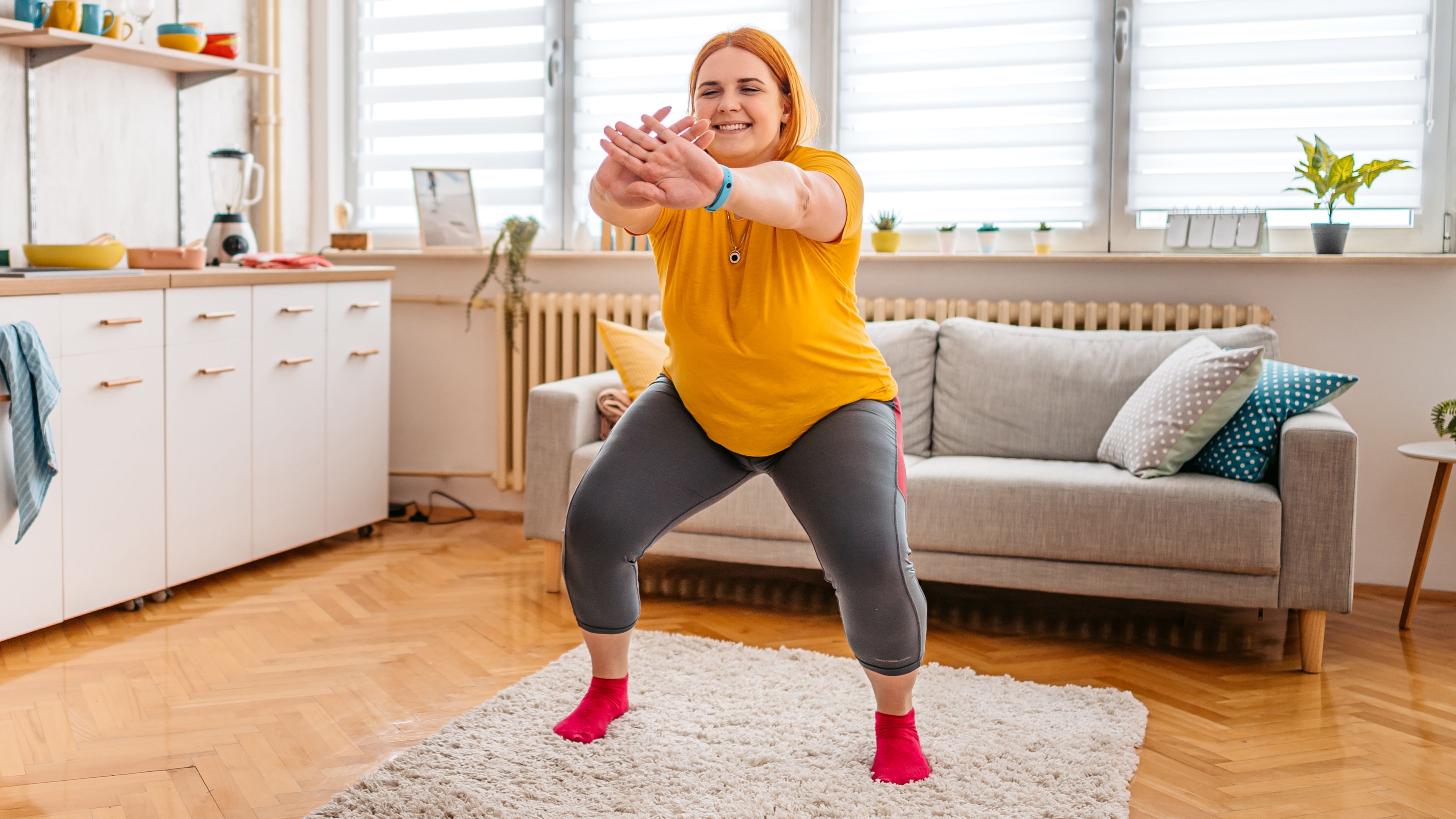 A woman wearing leggings and a t-shirt practices squats at home. She is standing on a rug, behind her we see a couch and to her side there is a kitchen unit with a sink and dishes. 