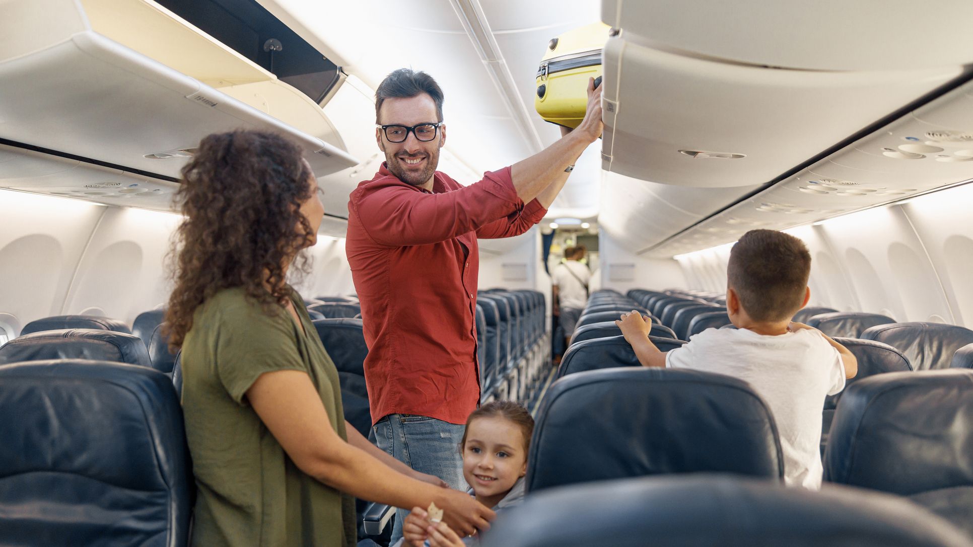Cheerful man looking at his wife with a smile and putting carry on luggage in compartment while traveling together with his family by plane