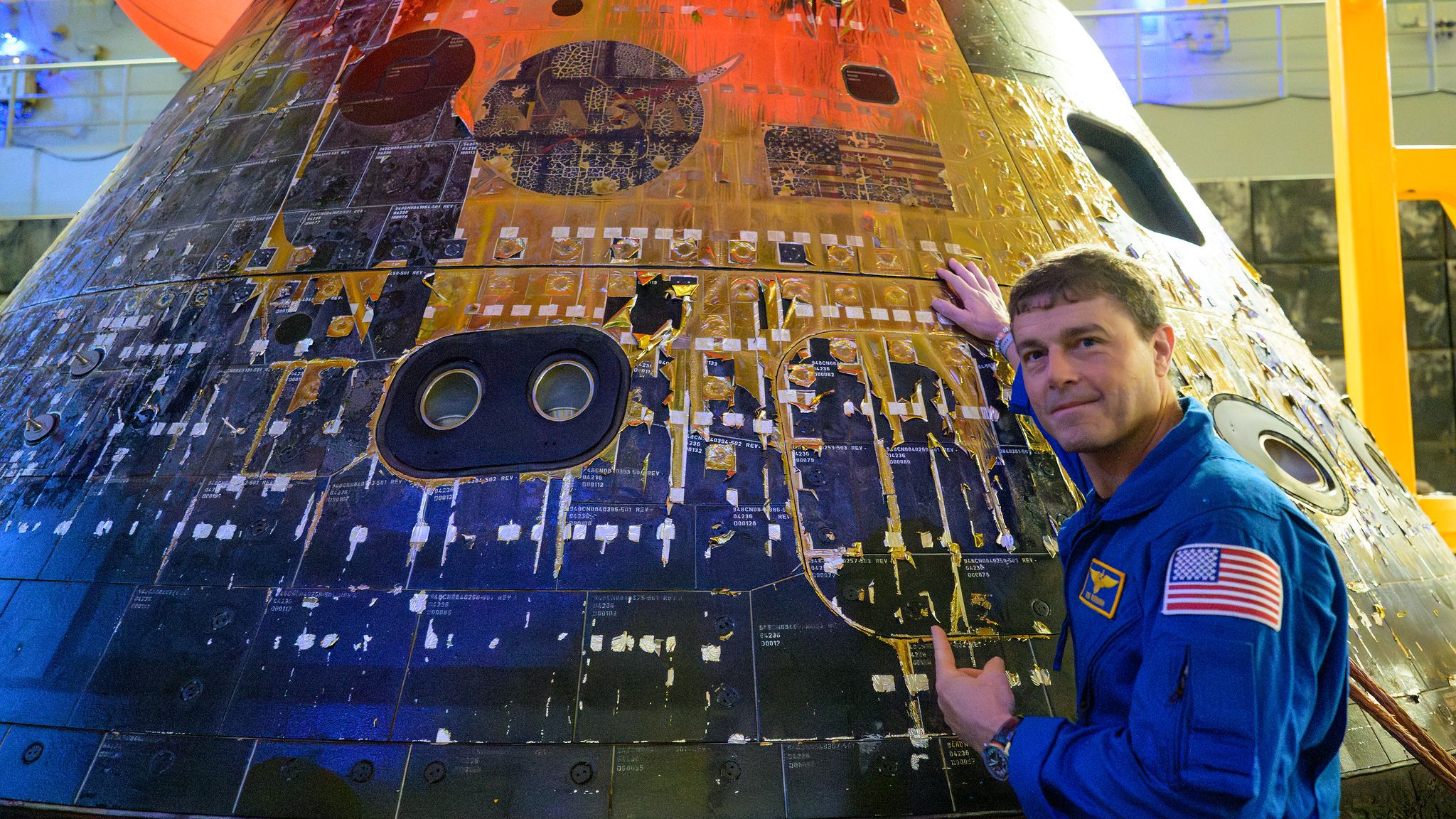 O comandante da Artemis 2, Reid Wiseman, é fotografado com um macacão azul da NASA olhando para a câmera com a mão na espaçonave Orion carbonizada enquanto ela está parada no convés de uma nave de recuperação.