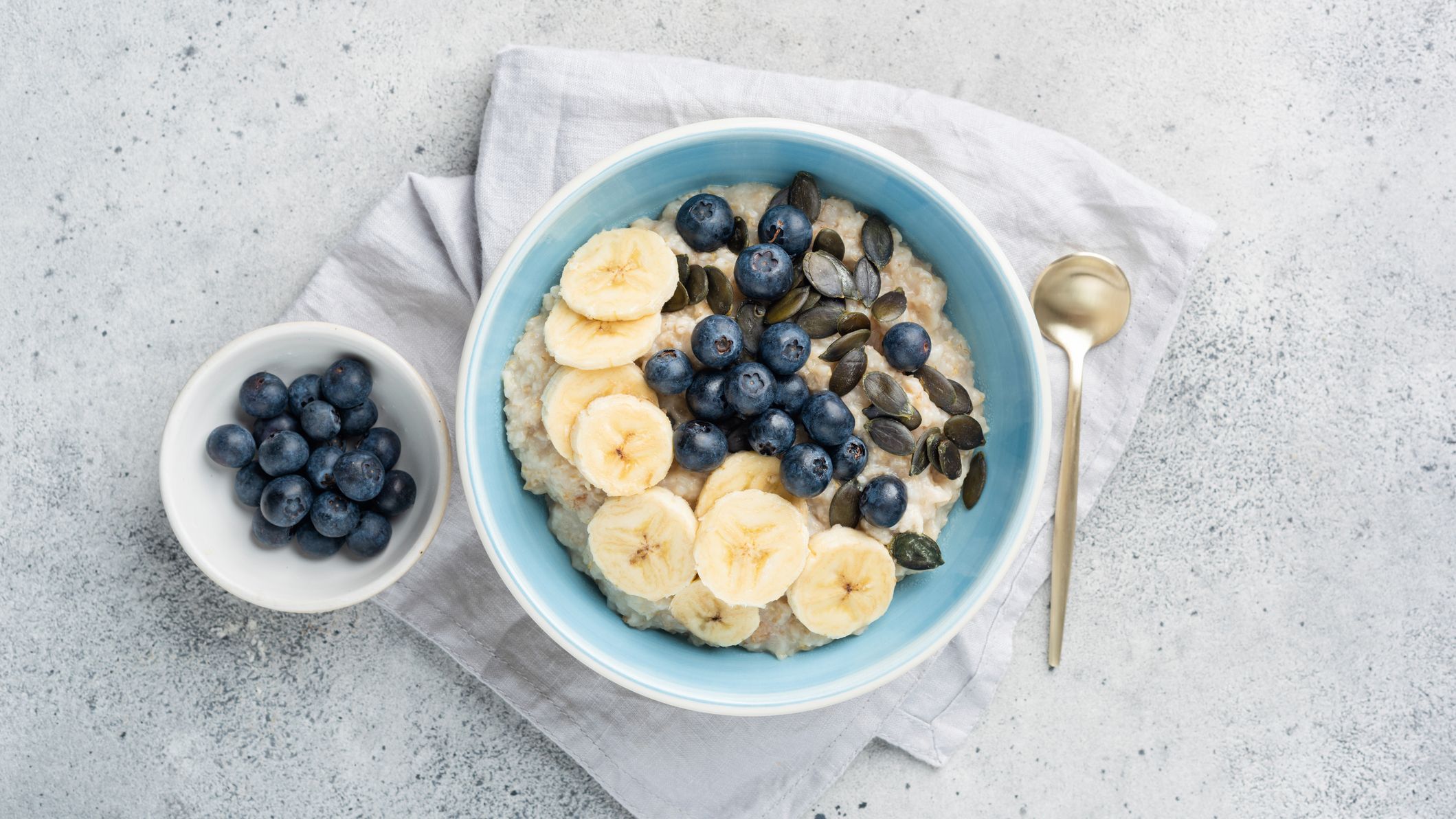 Bowl of porridge topped with slice banana, blueberries and pumpkin seeds. A smaller bowl of blueberries is to the left of the porridge bowl. Both bowls and a spoon rest on a white serviette.