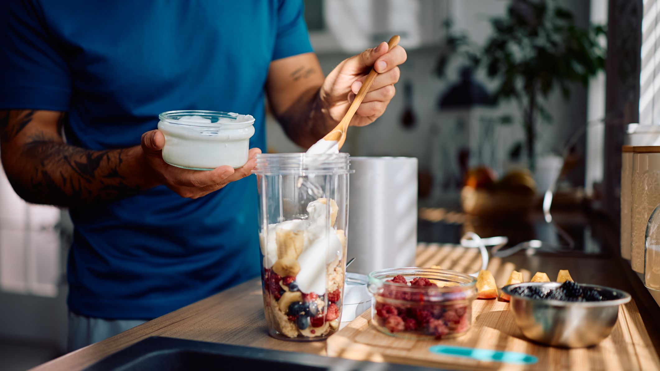 Close up of a man spooning yogurt into a blender jug resting on a counter, next to bowls of fruit