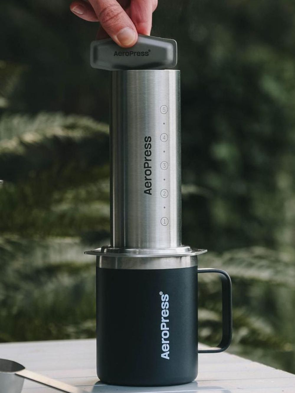 A stainless steel AeroPress tool on top of a black travel mug being held in place by someone's fingers on top, on a white table. Green leafy plants out of focus in the background.