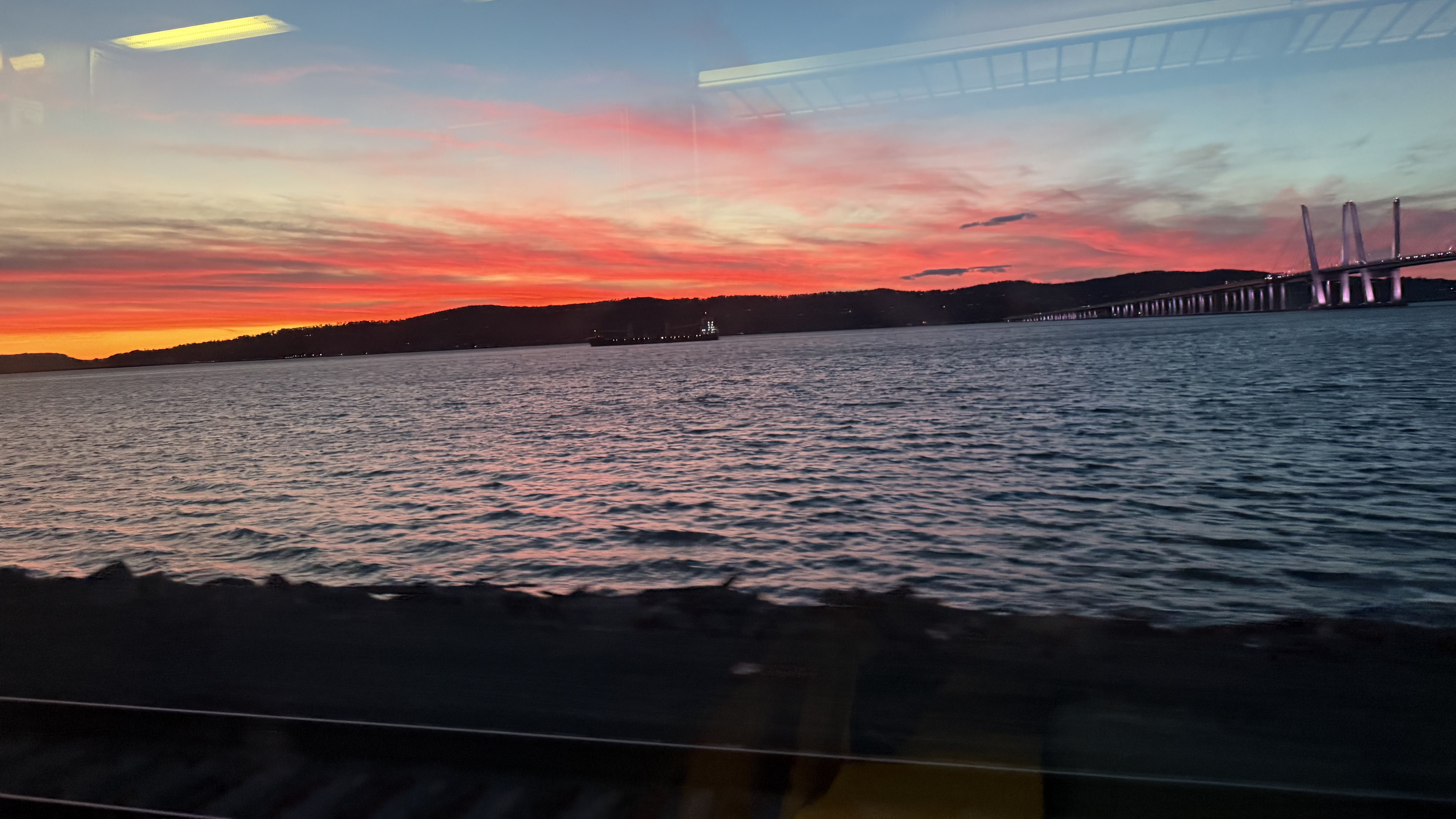 A colorful sunset over the cliffs with the Hudson River and train tracks in the foreground, and a bridge to the right.