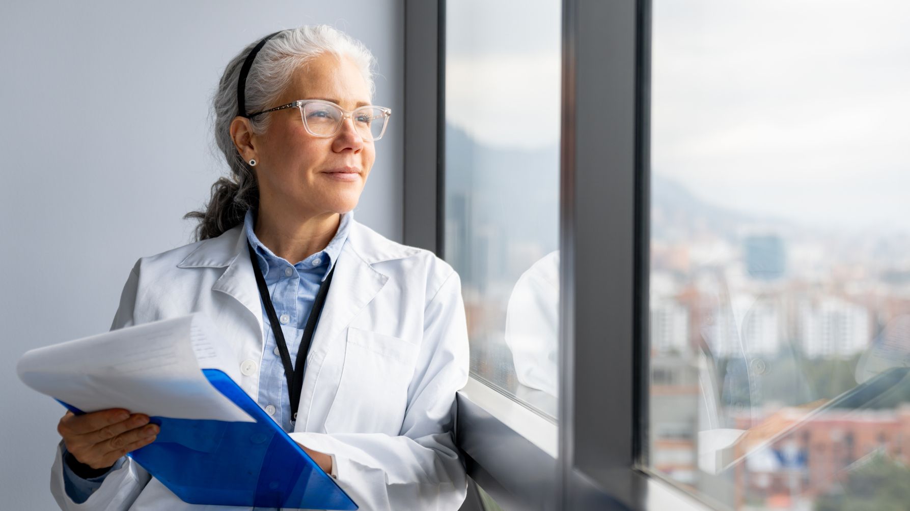 A female doctor holding a clipboard at the hospital and smiling while looking through the window.