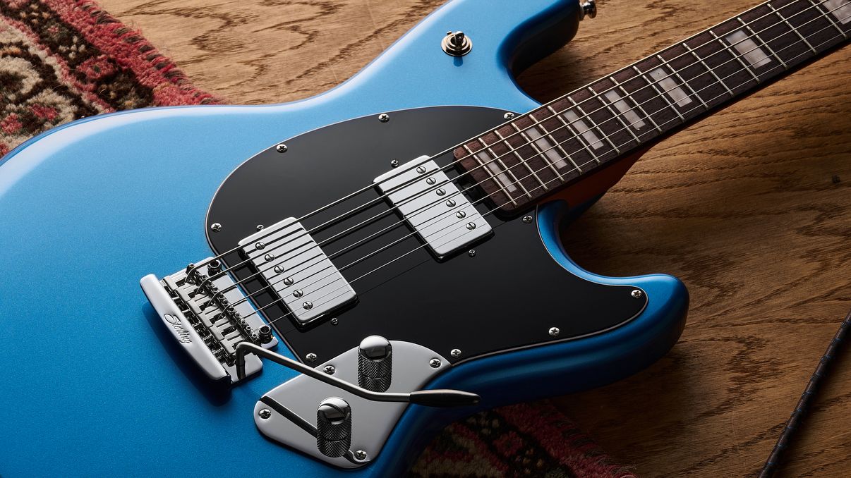 A Sterling by Music Man StingRay Baritone guitar lying half on a rug and half on a wooden floor. A guitar cable is trailed nearby