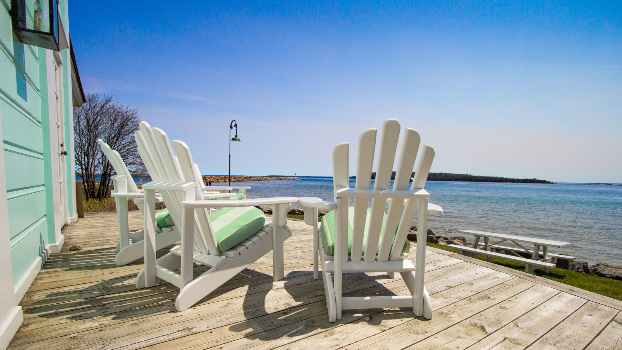 Three white chairs sit empty on a house's deck overlooking the ocean or beach.