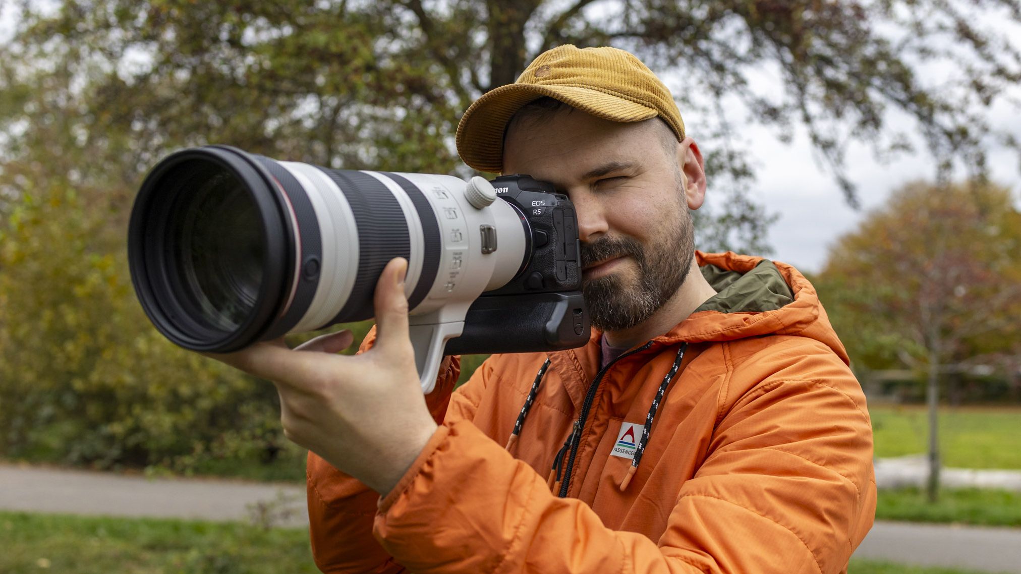 Photographer Dan Mold outdoors wearing an orange waterproof jacket and a yellow cap, holding a Canon EOS R5 with the Canon RF 100-300mm f2.8 lens mounted to it up to his eye