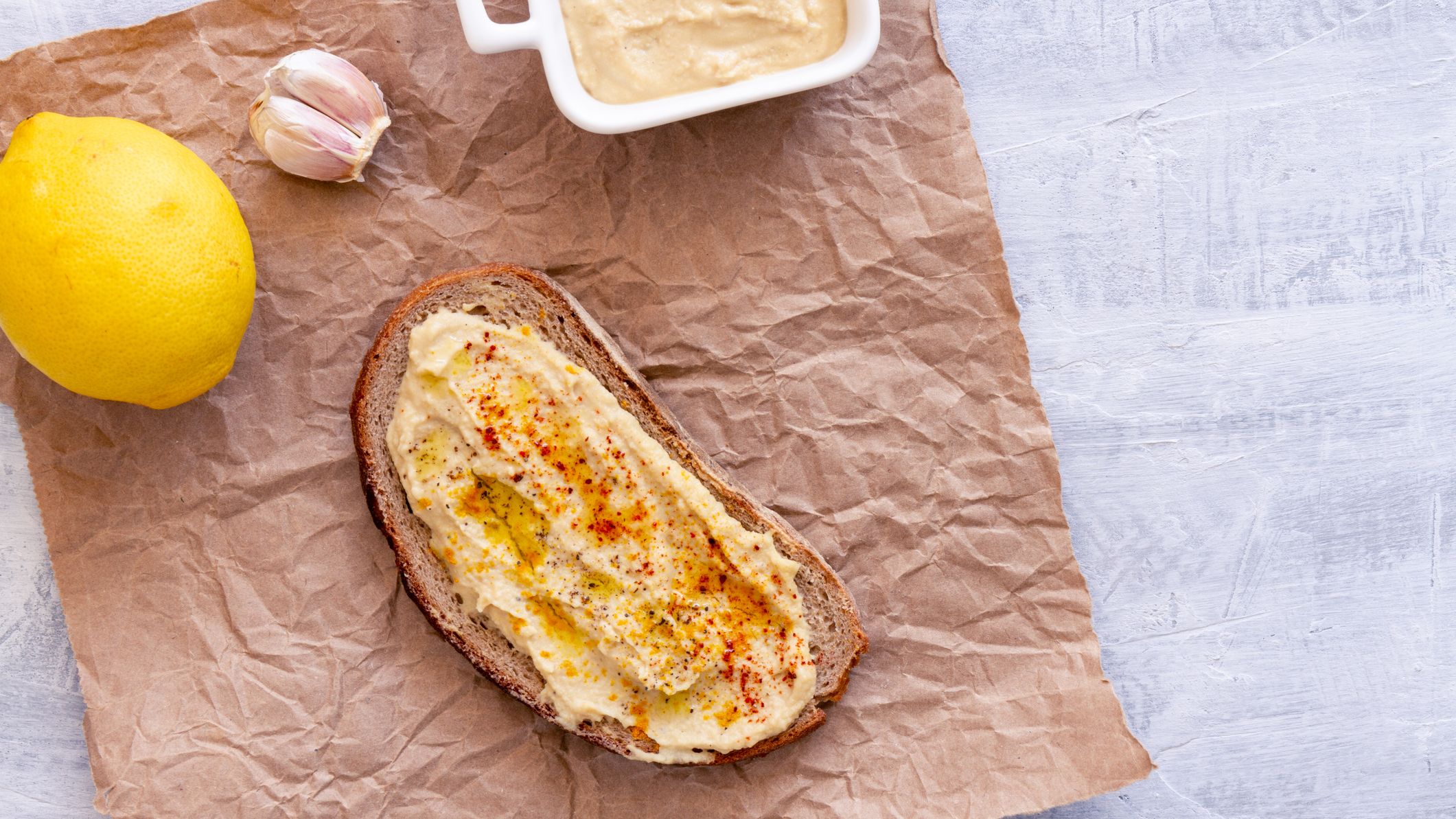 Hummus spread on toast seen, a lemon, two unpeeled garlic cloves and a small container of hummus seen from above,
