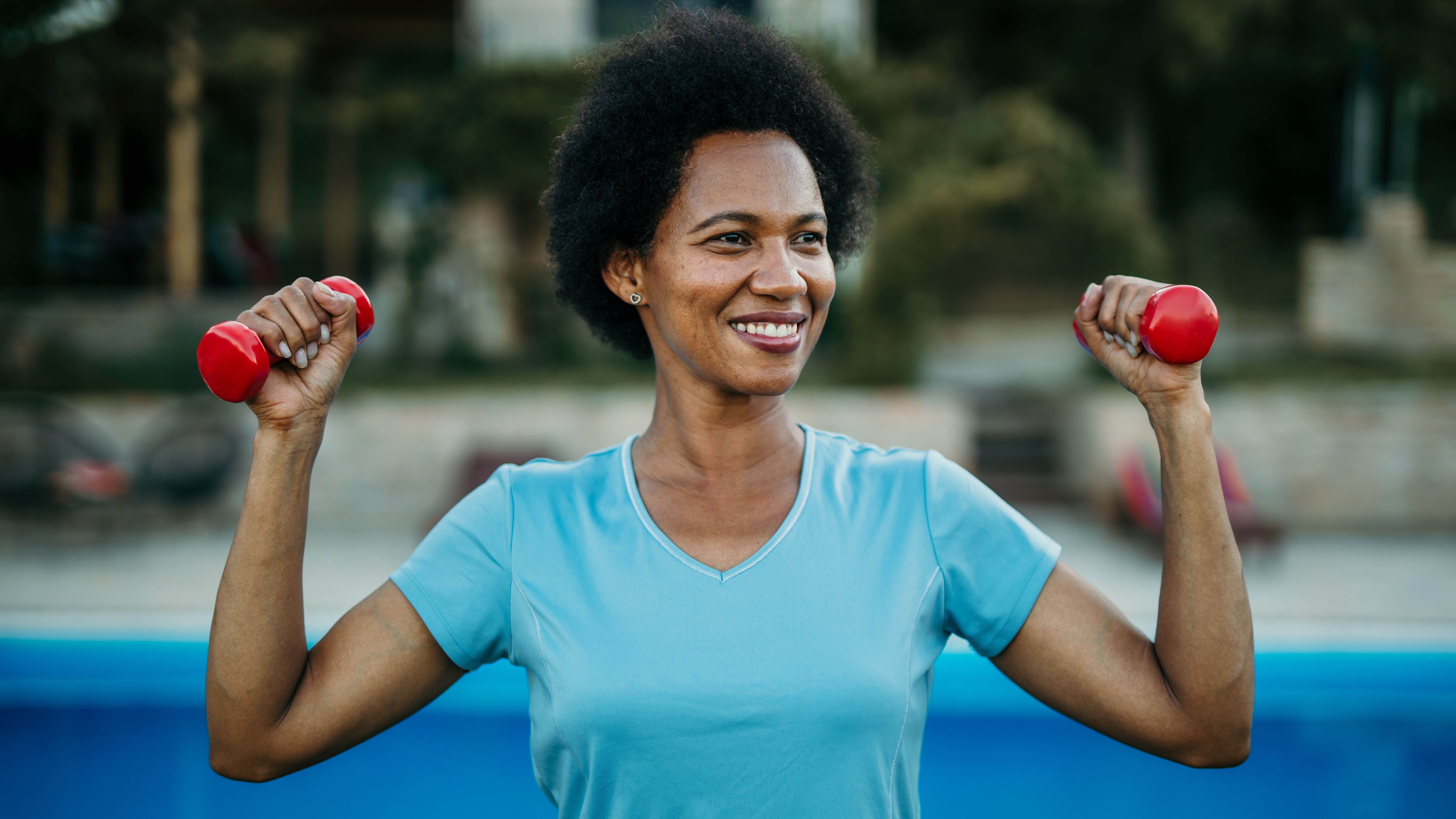 A woman stands outside holding a pair of light dumbbells in her hands at shoulder height and smiling.