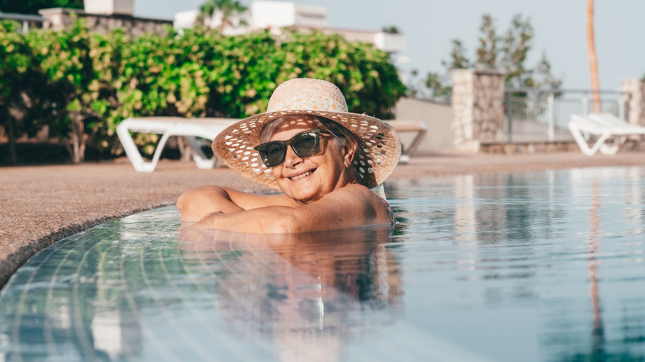 Woman with hat and sunglasses relaxing in an outdoor swimming pool at a resort.