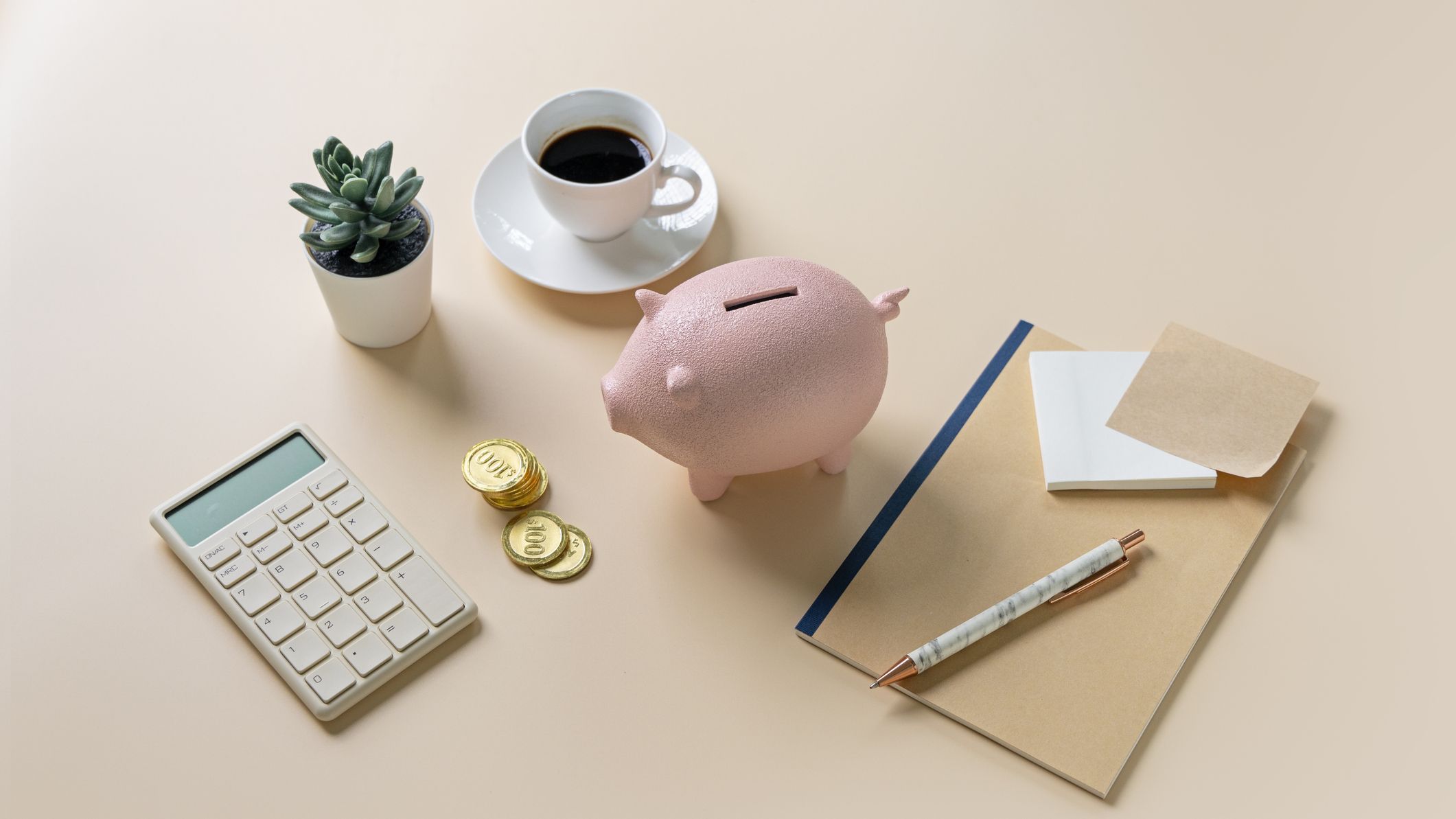 a desk with a notepad, calculator, small plant, a cup of coffee and a piggy bank