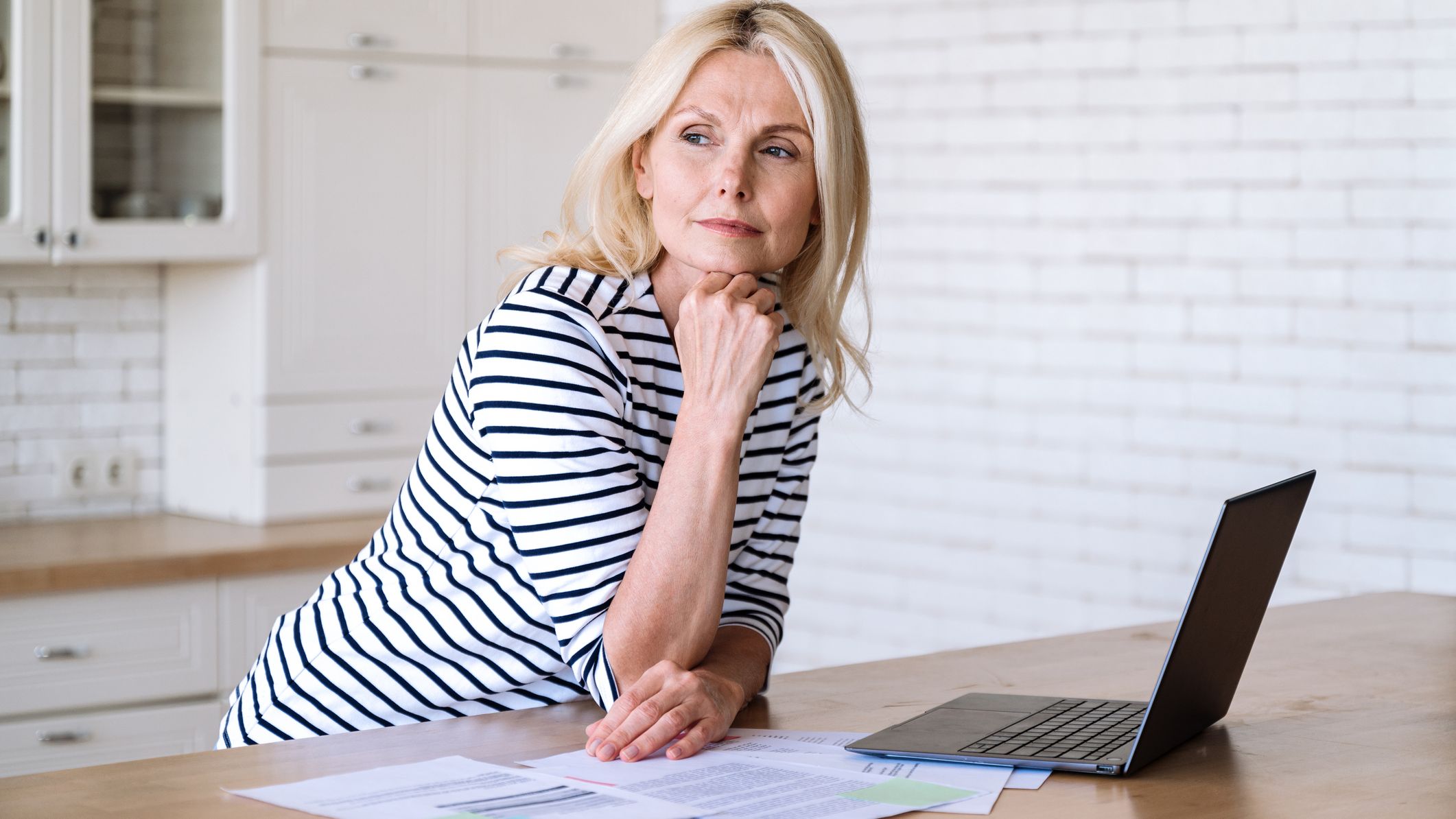 Thoughtful female standing with laptop and documents on desk