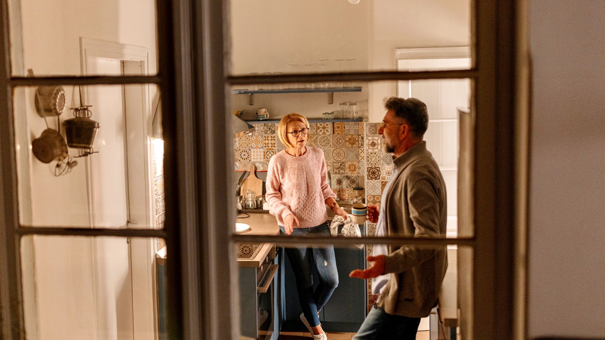 View of a senior couple in their kitchen having an argument. The viewer is looking through the window from outside the house.