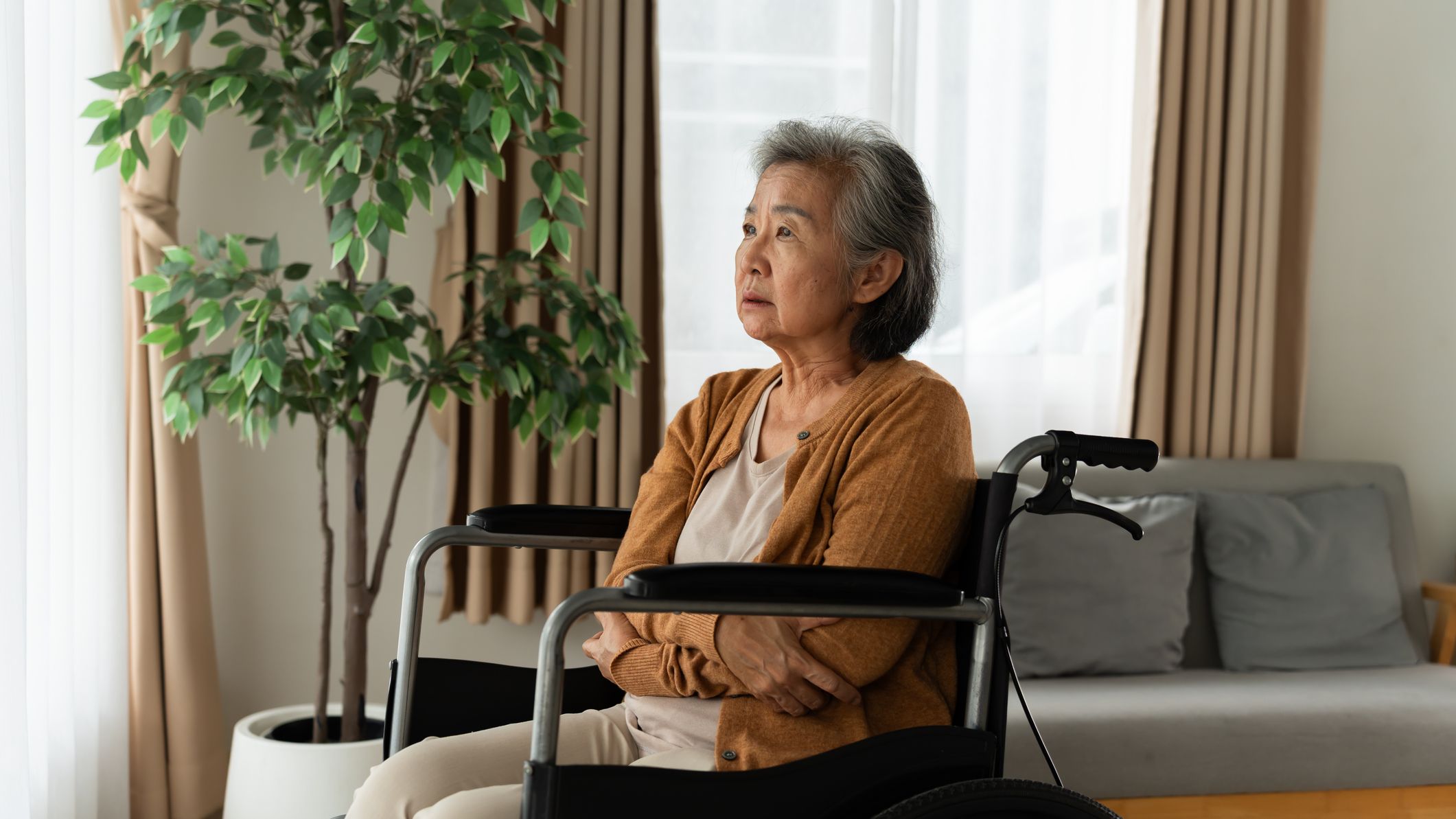 A woman sitting in a wheelchair at home and looking toward a bright window.