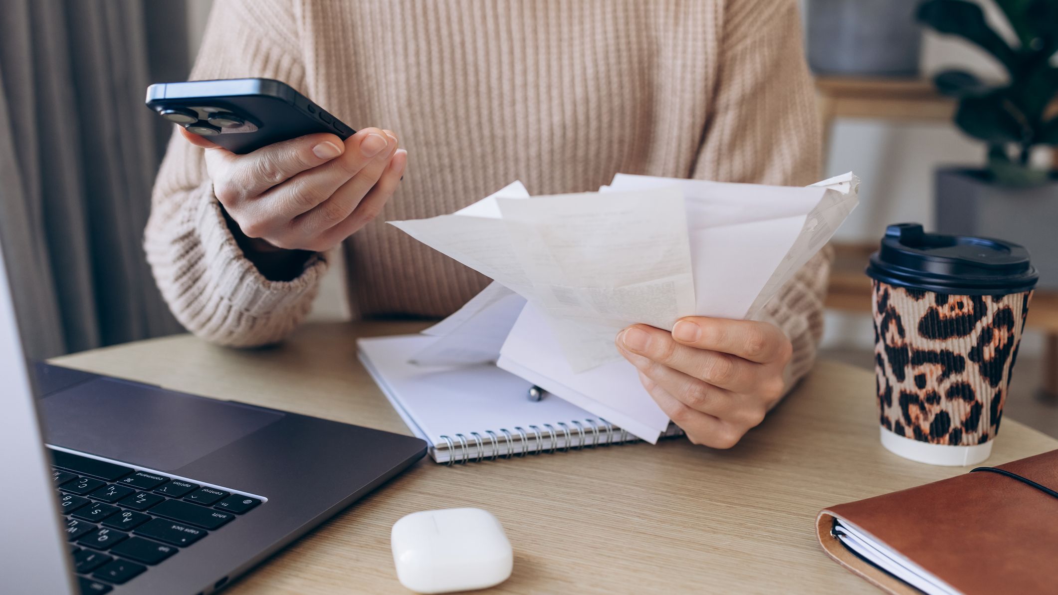 Close-up of a woman reviewing receipts while holding a smartphone beside an open laptop at a cozy desk.