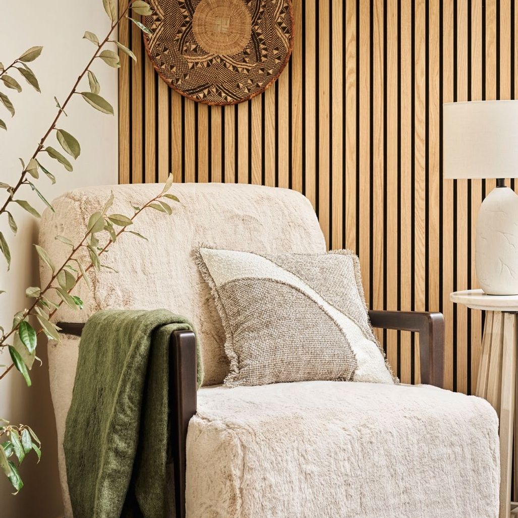 White armchair in teddy material, green throw draped over the arm with a side table and lamp to the side with acoustic wall panelling in the background.