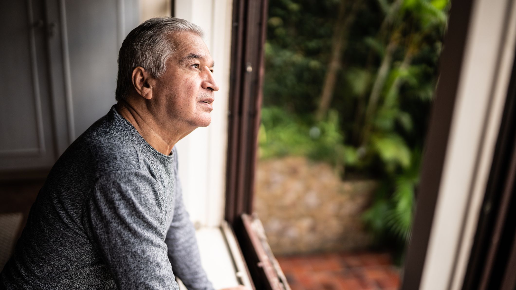 An older man is contemplating while looking through a window to the outdoors.