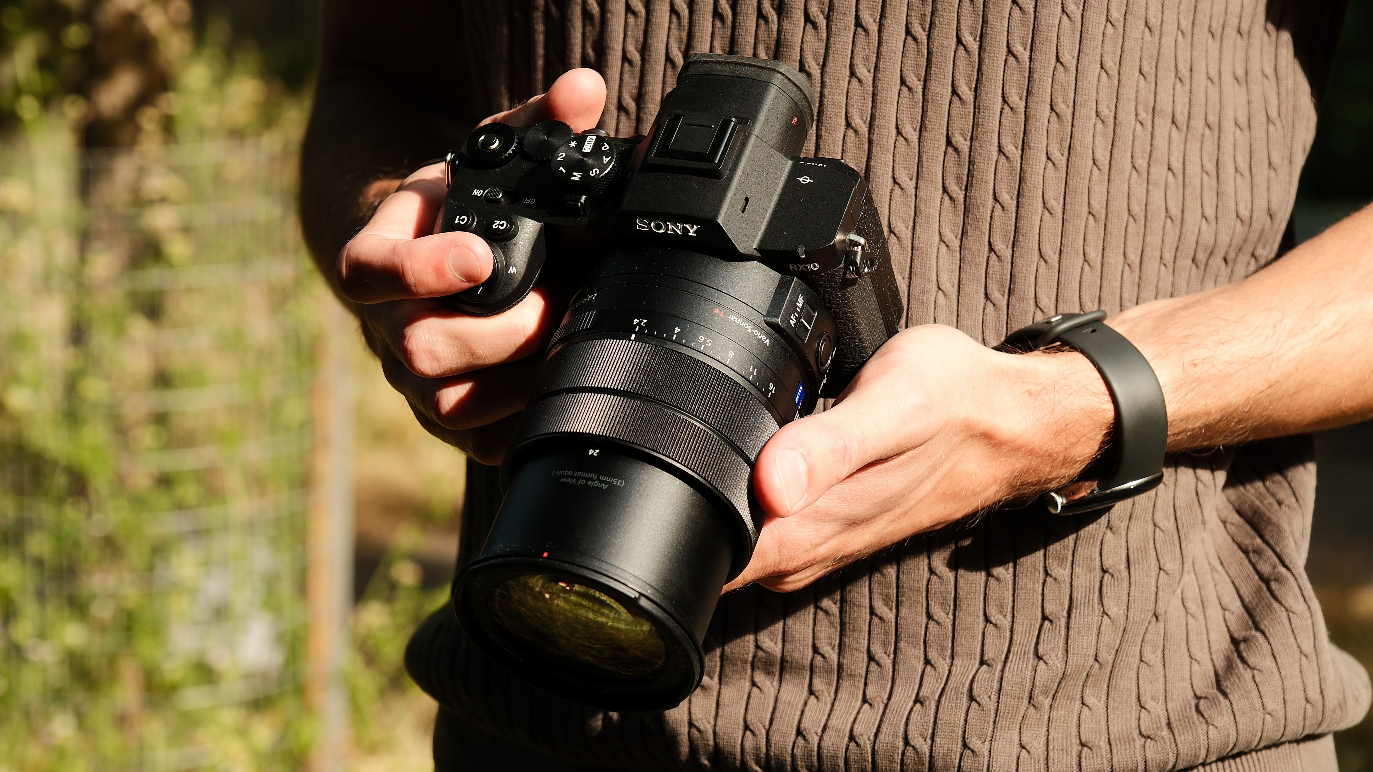 Person holding the Sony RX10 V with both hands outdoors in bright sunlight.