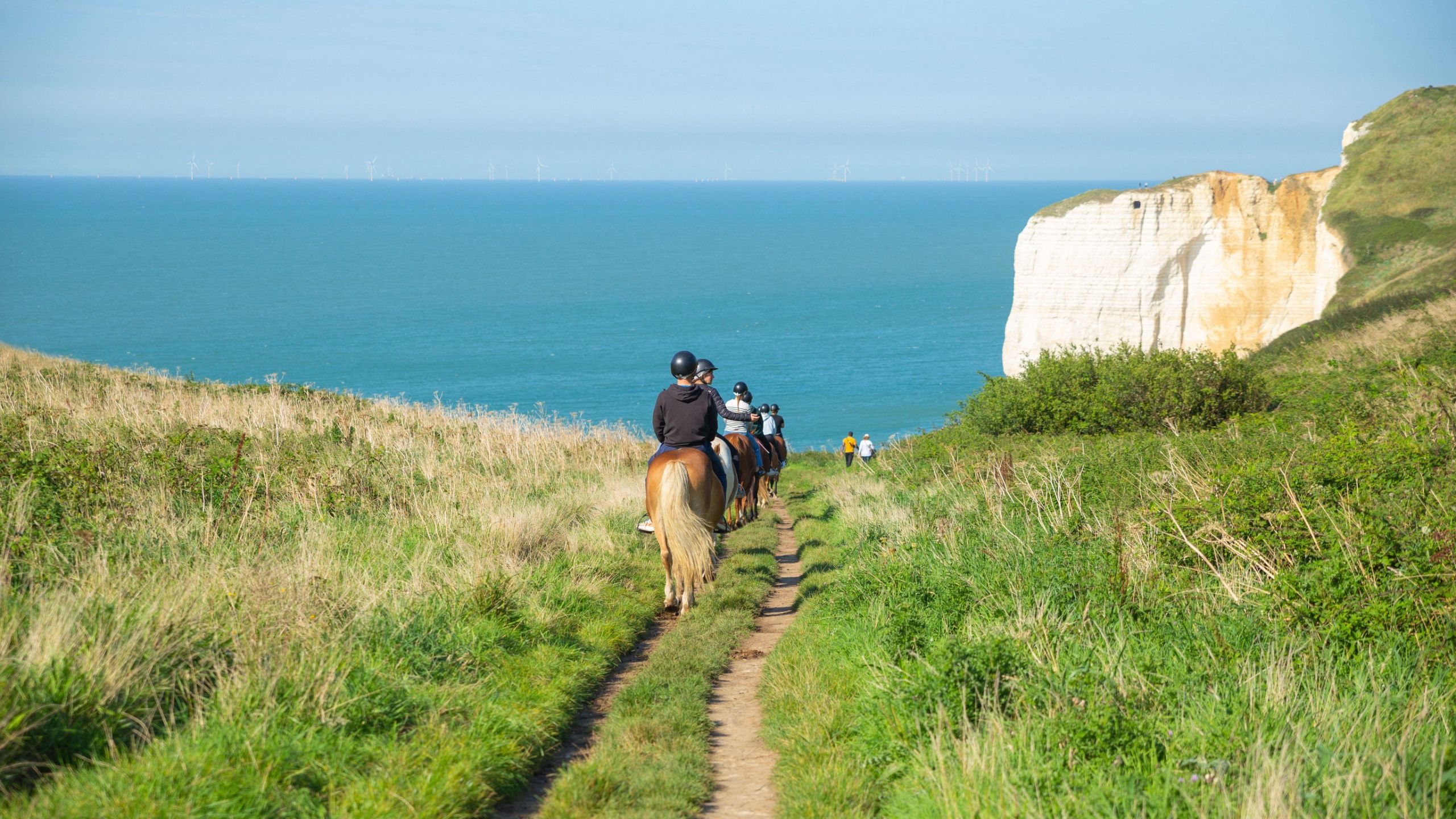 a group of riders pictured in single file from behind, descending a coastal path. There is blue sea and a white cliff protruding into it in the distance