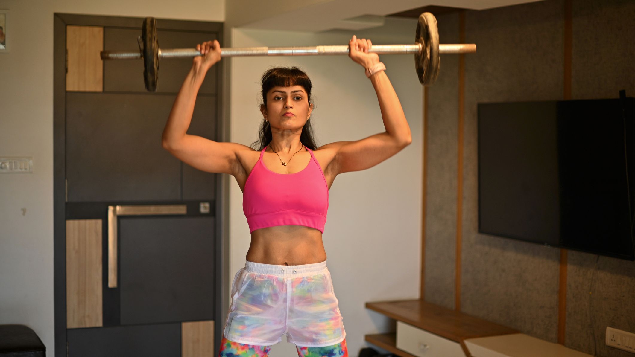 Woman lifts small barbell above her head in a domestic setting