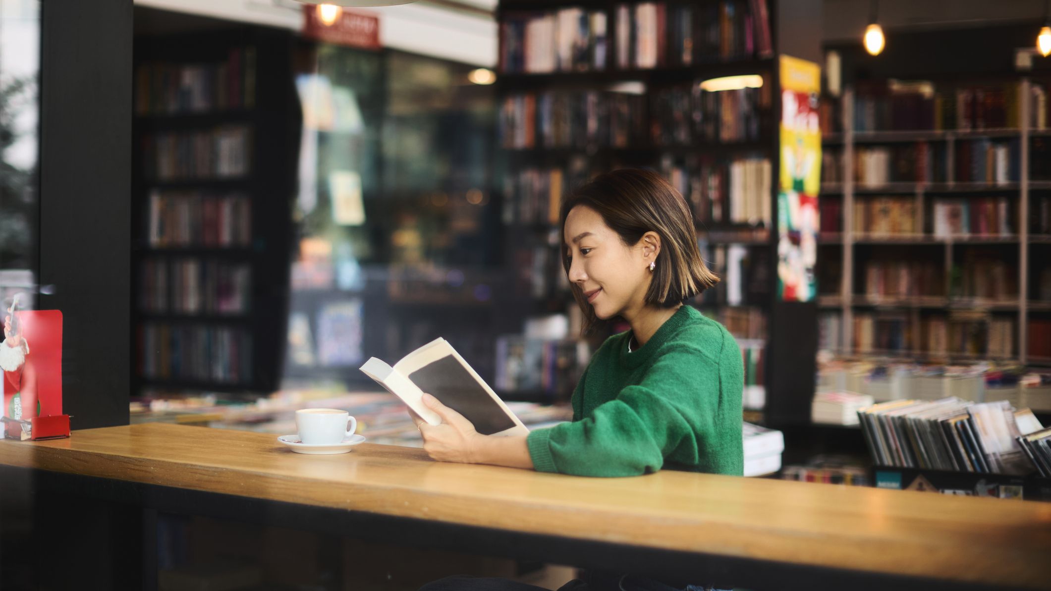 A woman sits in a cafe by the window and reads a book with a cup of coffee