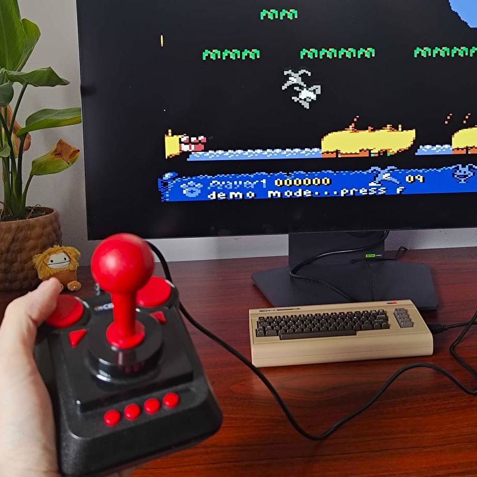 Hand holding C64 Mini joystick with C64 Mini in backdrop connected to monitor displaying Nobby the Aardvark gameplay on screen.