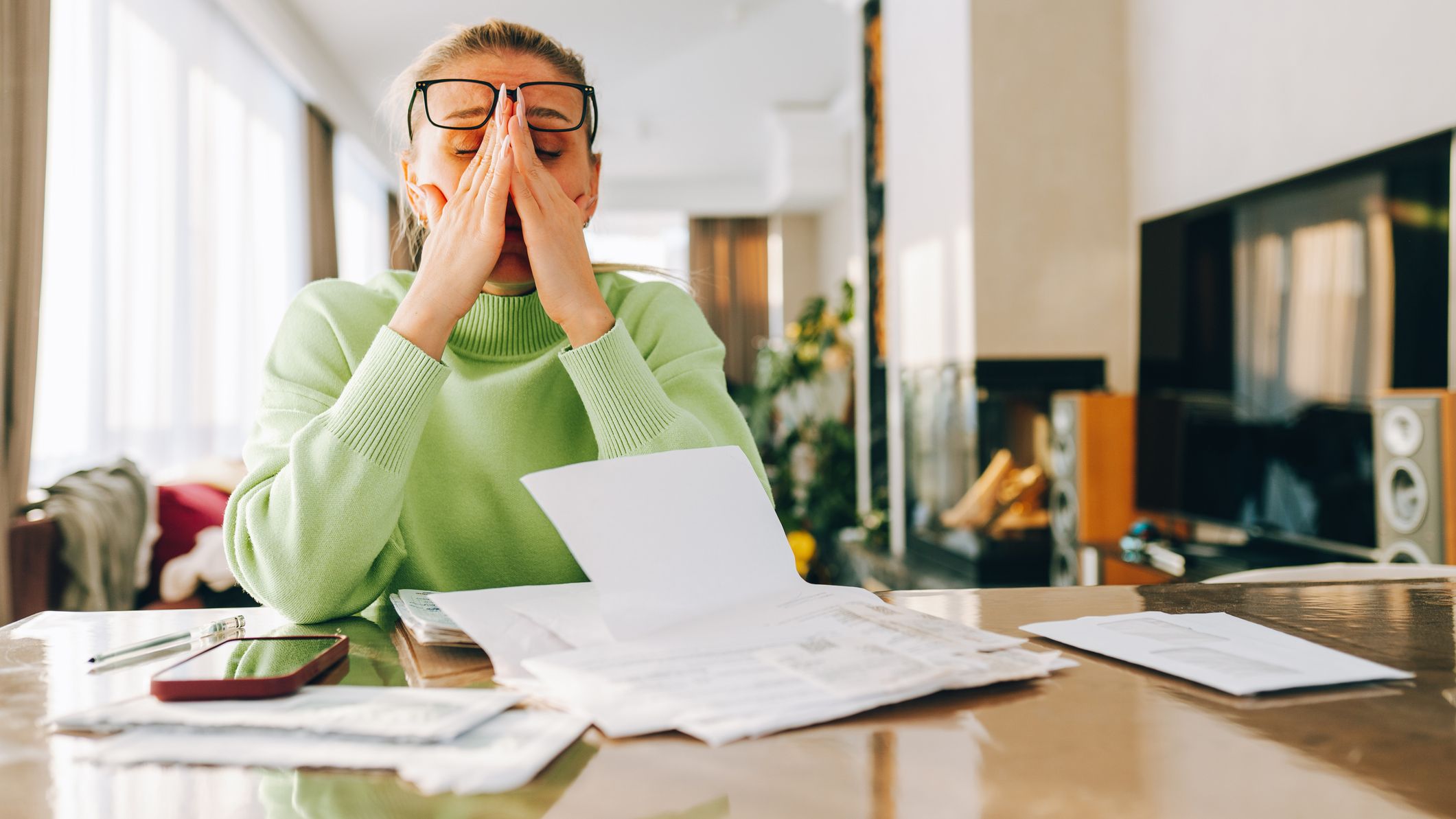 A stressed woman rubs her temple while reviewing financial paperwork.