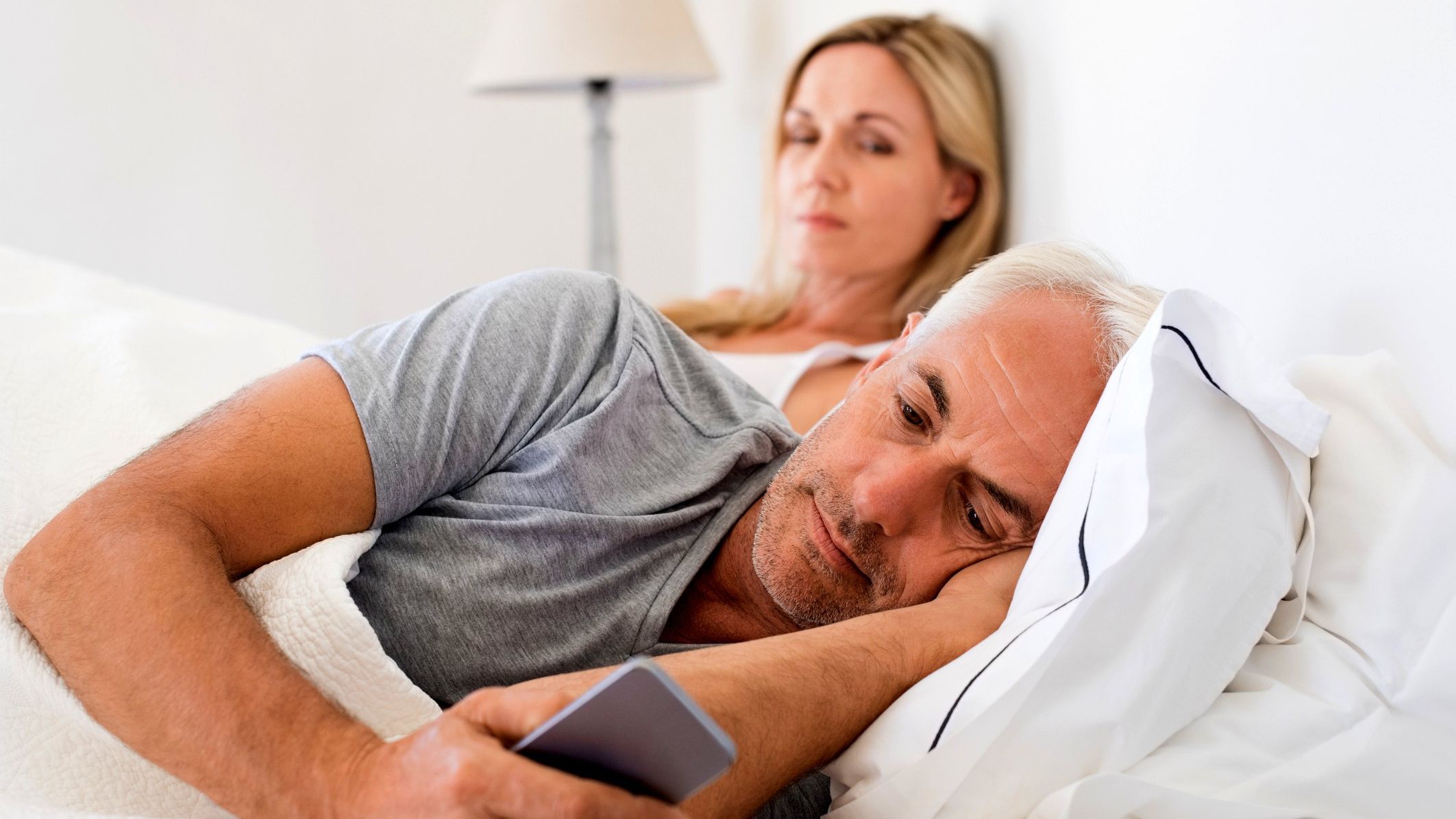 A couple, perhaps in their 50s, rests in bed. The man is looking at his cell phone and turned away from his wife or partner.