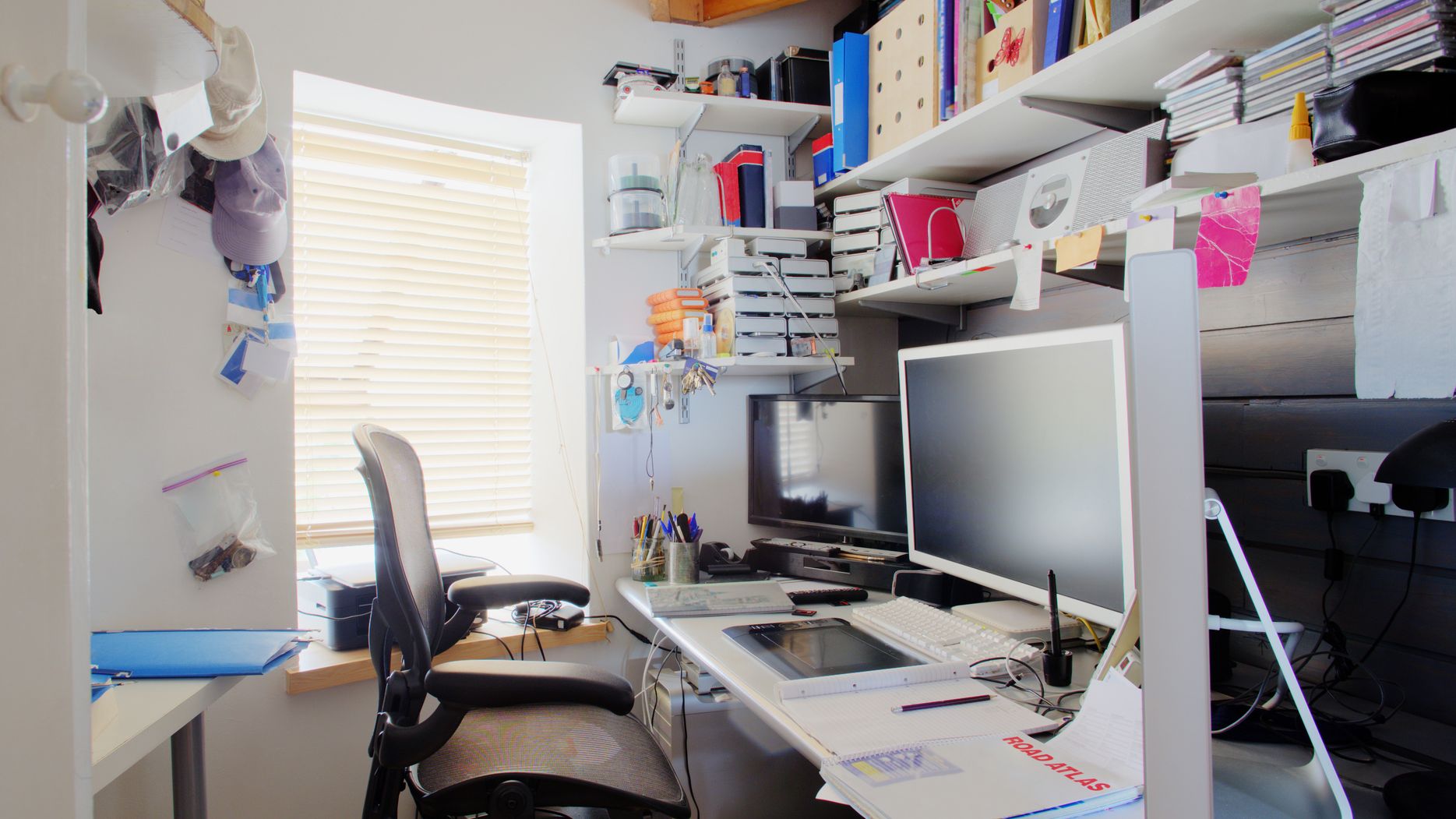A shot of a messy desk in a home office, the room is small and cluttered, on the desk is three computer monitors and office supplies