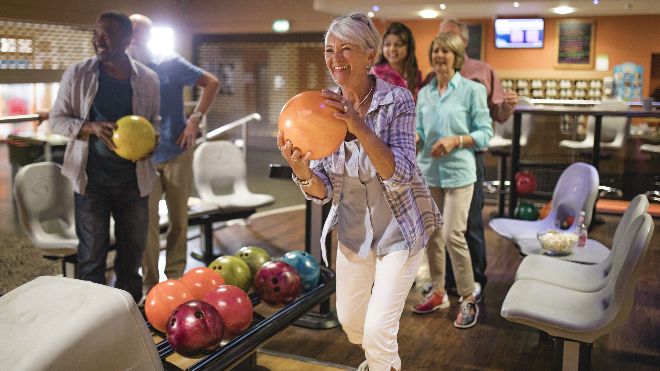 Senior woman holding bowling bowl about to bowl in bowling alley. A group of people stand behind her watching on