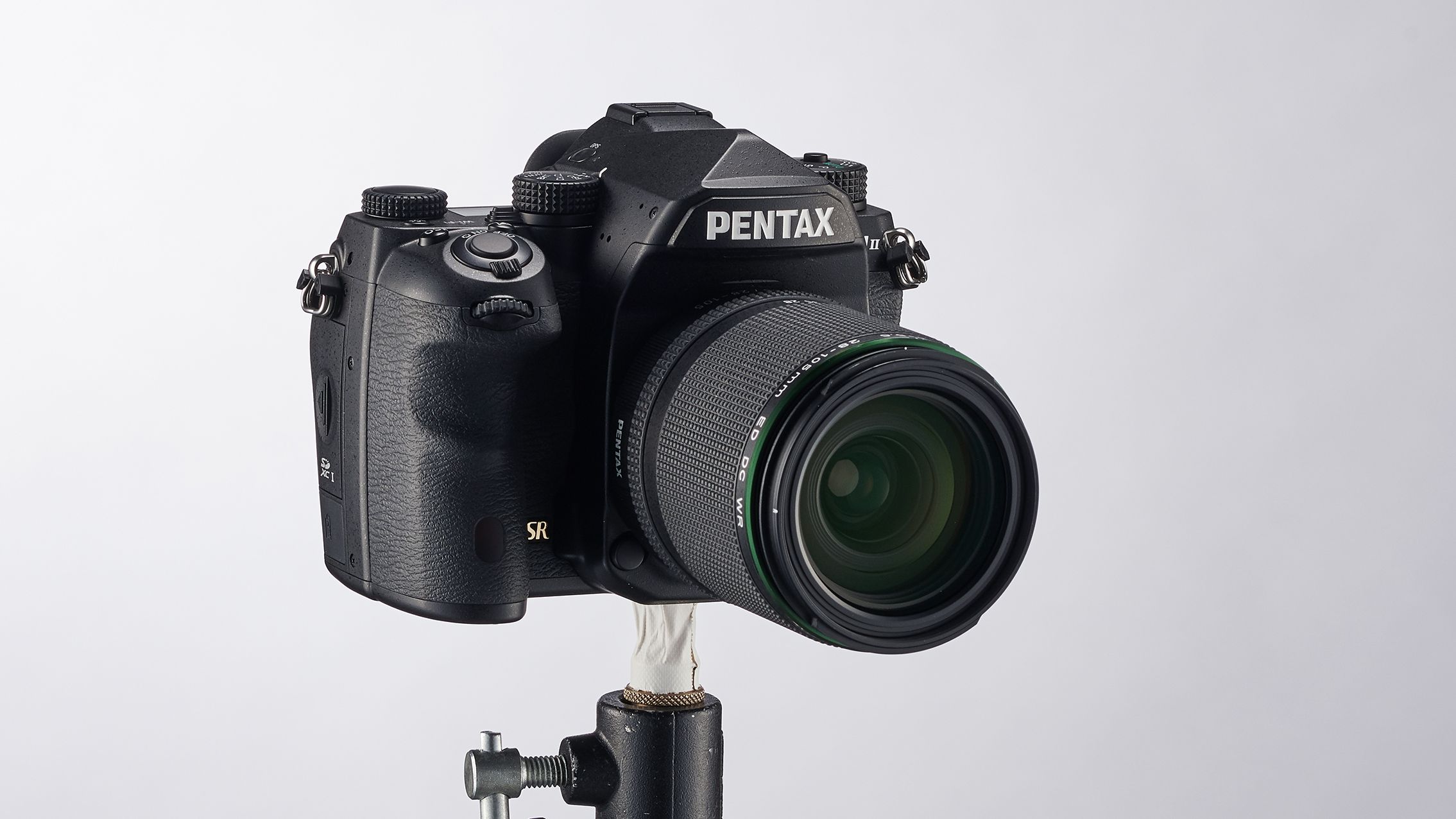 Black Pentax K-1 Mark II DSLR camera with zoom lens mounted on a small metal clamp against a plain white background