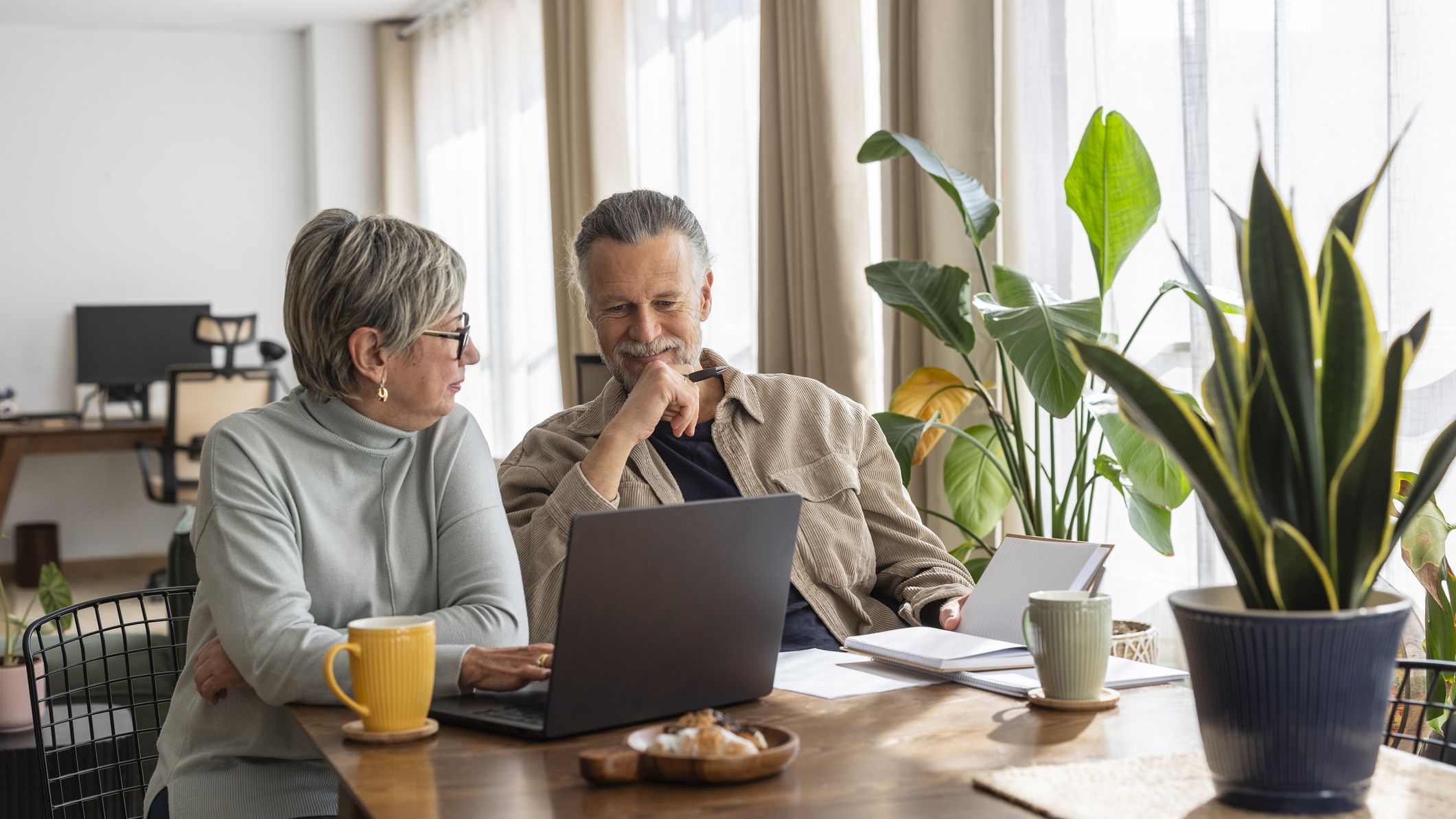 A couple in their 60s discusses finances or other planning in front of a laptop. They sit at the dining room table with coffee, food and light coming through the windows.