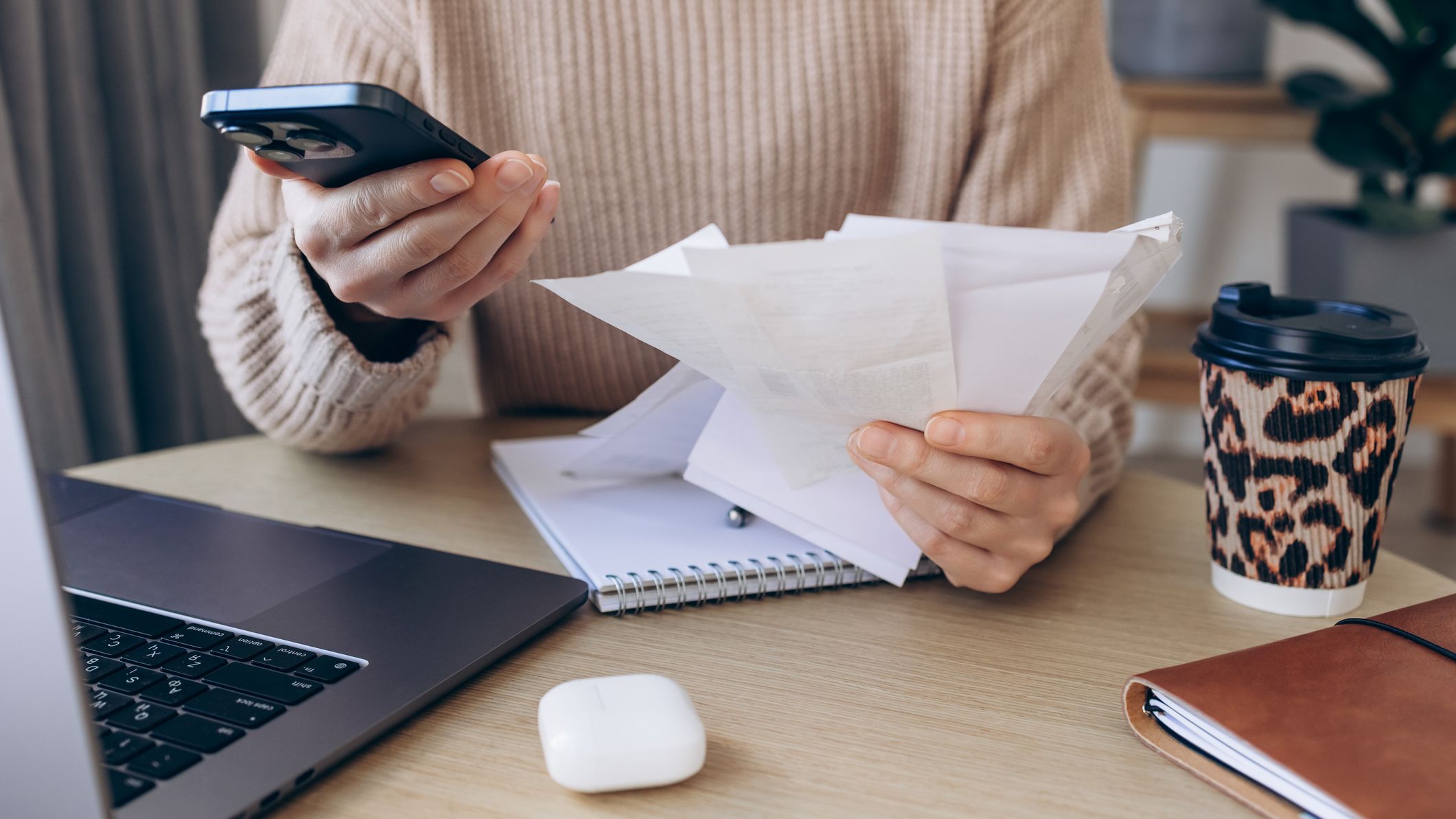 Close-up of a woman reviewing receipts while holding a smartphone beside an open laptop at a cozy desk.