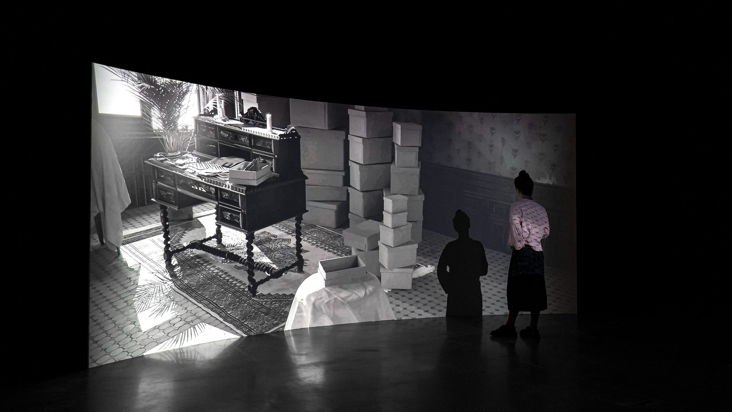 A curved screen in a dark gallery shows six identical images of a woman in glasses, with Spanish subtitles below