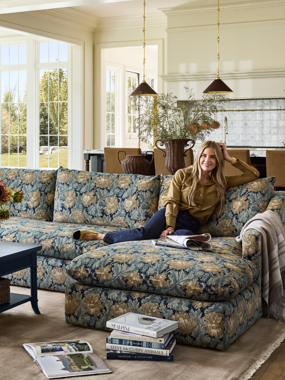 Shea McGee smiles on a floral jacquard sectional from her new McGee &amp;amp; Co. fall 2026 collection, which is styled with a navy storage coffee table, taupe rug, woven table lamp, and collected objects