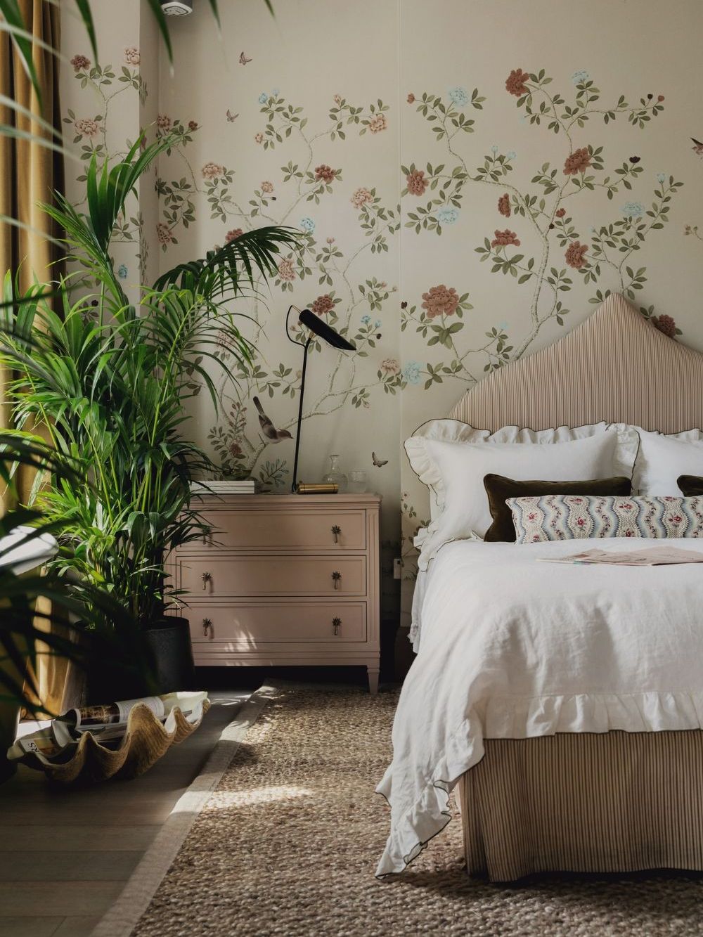 soft pink bedroom with floral mural wallpaper, a pink striped headboard, pink nightstand and white ruffle trimmed bedding with house plants