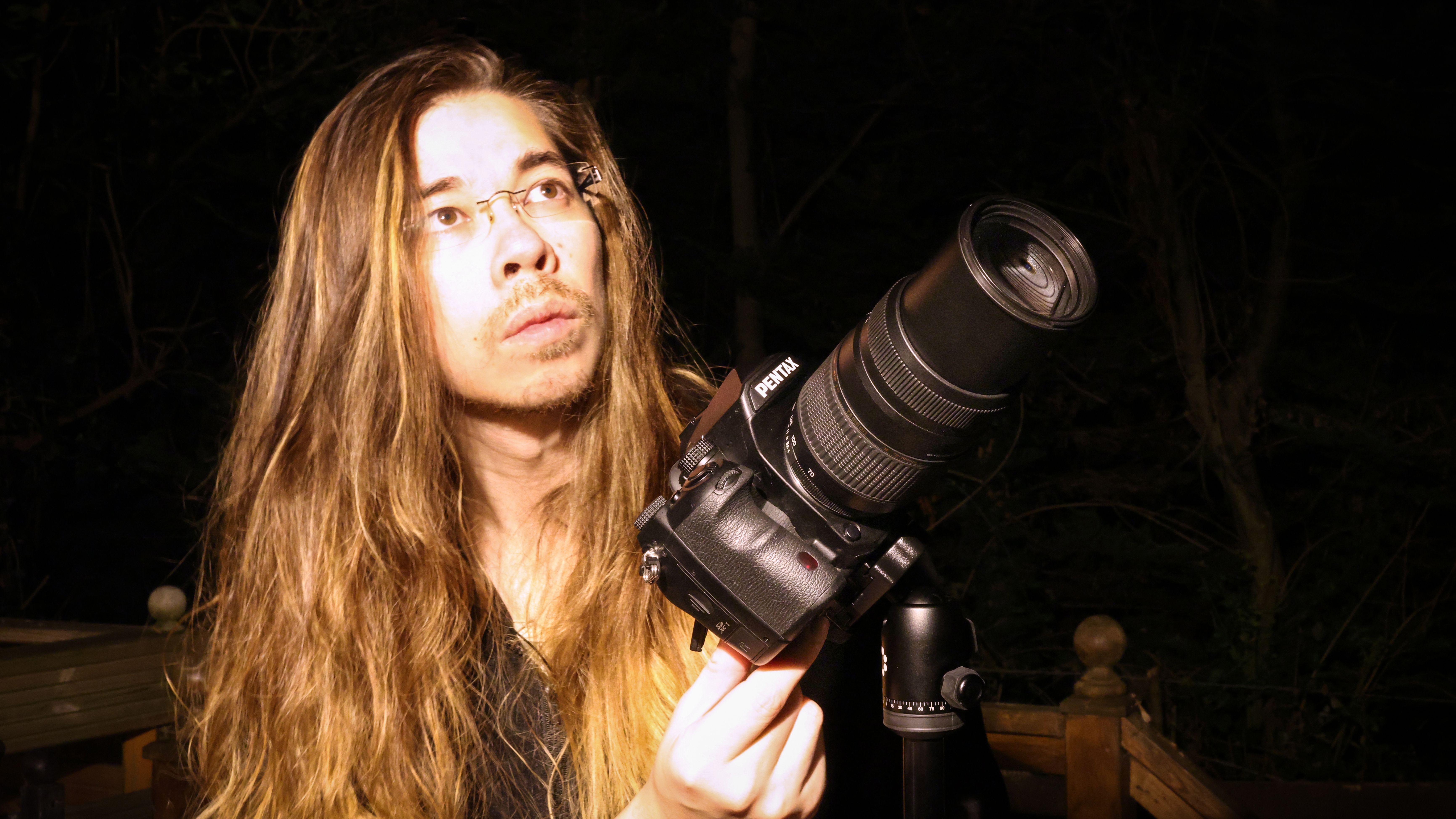 Man with long hair holding a Pentax K-1 Mark II at night
