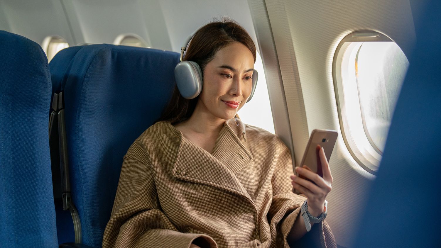 A chic woman is seen relaxing at the window seat on a flight, enjoying her time with wireless headphones and a smartphone