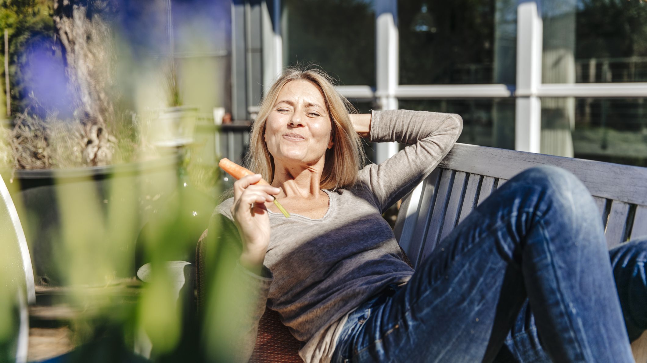 A woman in her late 40s or early 50s relaxing on a garden bench, eating a carrot.