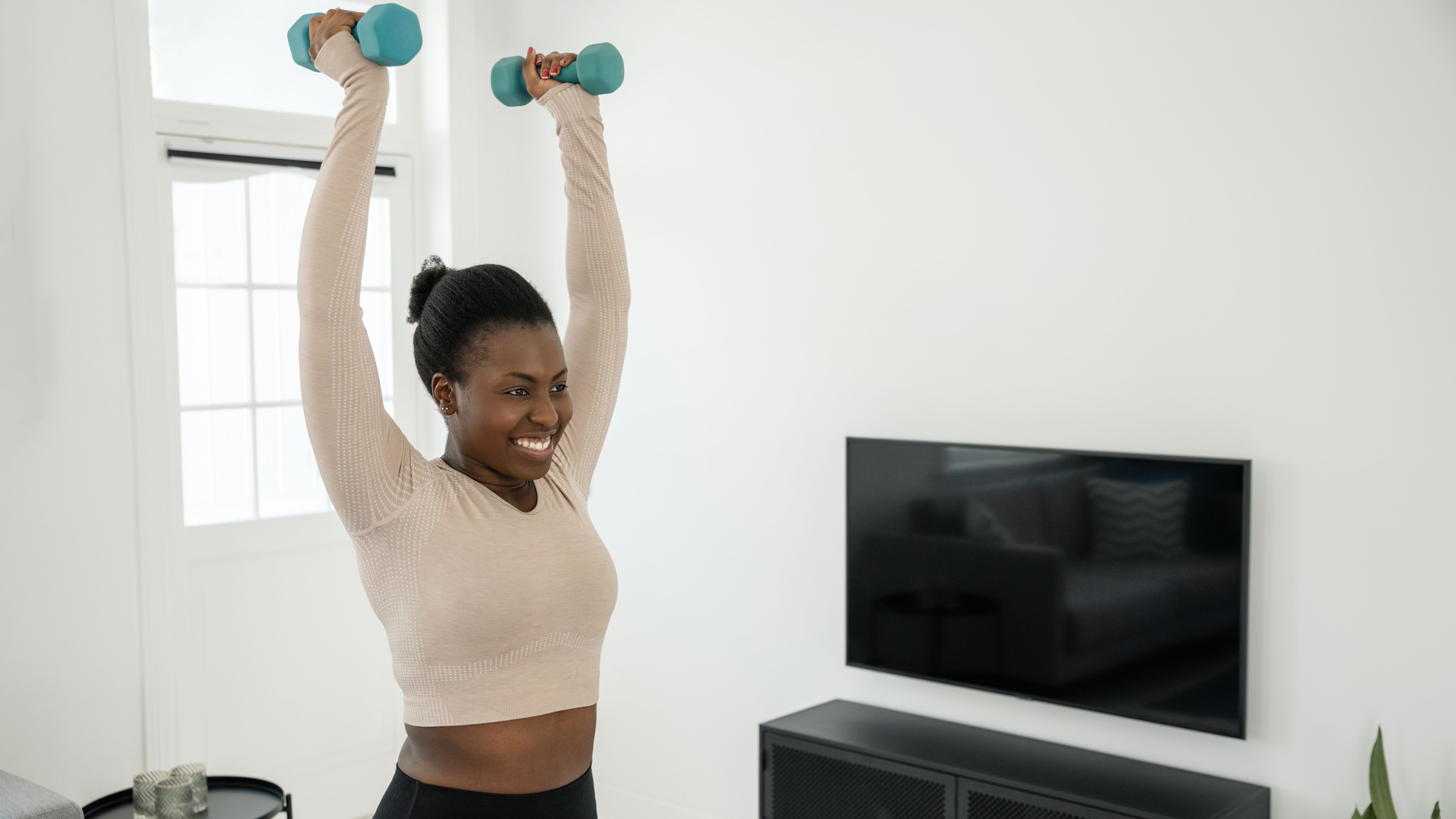 A smiling woman in long-sleeved crop top and leggings stands holding a pair of dumbbells overhead in her living room. Behind her we see a TV, plant and table.
