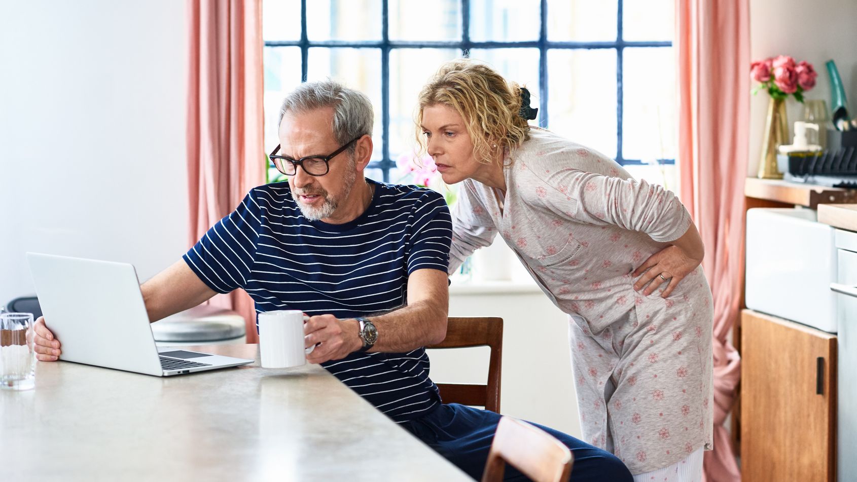 A mature couple in their kitchen look concerned while reviewing finances on their laptop.