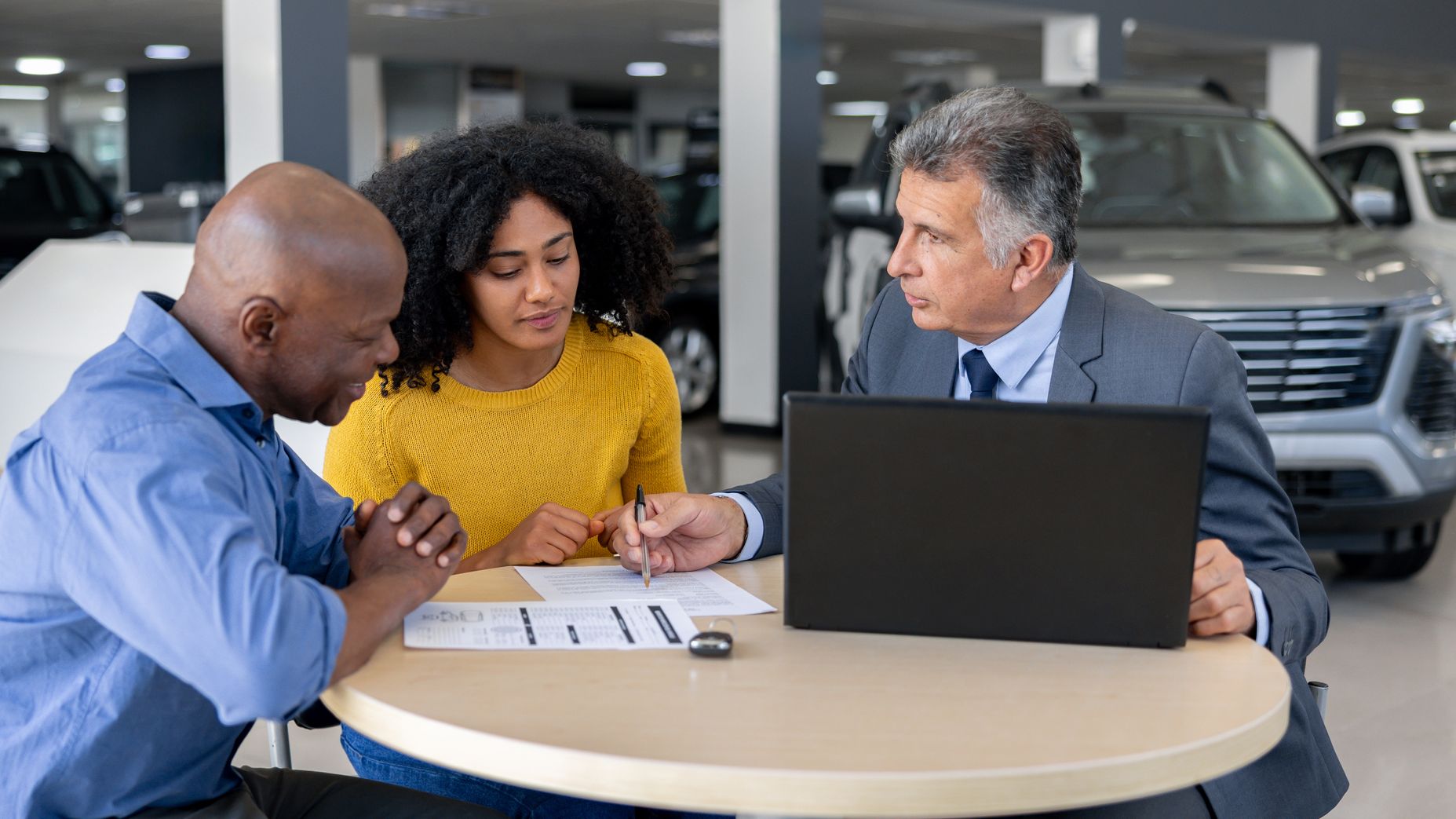 A car salesman discusses a contract with potential buyers while seated at a table inside a dealership.