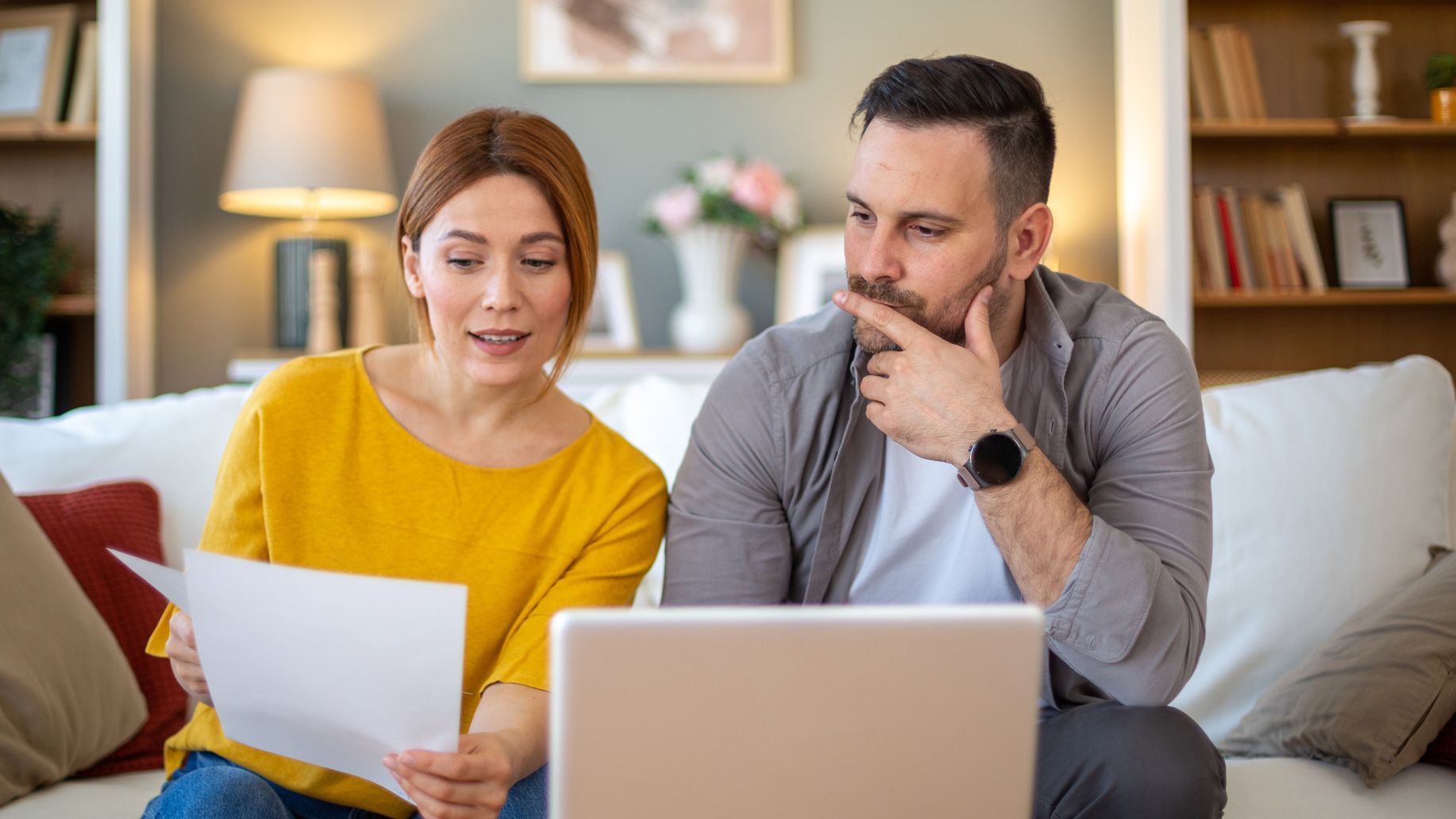 Married couple is sitting on sofa at home in front of laptop, discussing home finances.